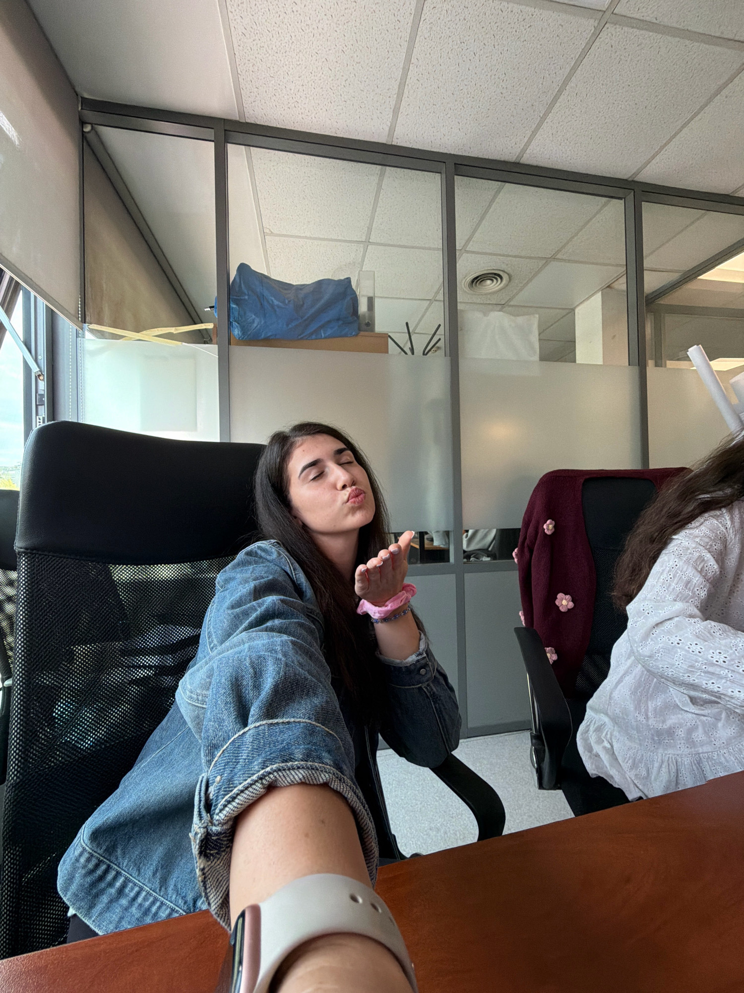 An indoor selfie is taken by a young woman sitting in a black office chair. She has long, dark hair and is wearing a denim jacket. She is puckering her lips as if blowing a kiss and her right hand is extended towards the camera, wearing a pink bracelet. Behind her, another person is partially visible wearing a white, textured top. The environment appears to be an office with glass partition walls, a suspended ceiling, and blinds covering a window. A desk is in the foreground, and a dark red cardigan with floral embellishments is draped over the other office chair. The scene takes place in an office setting in Sofia, Bulgaria.