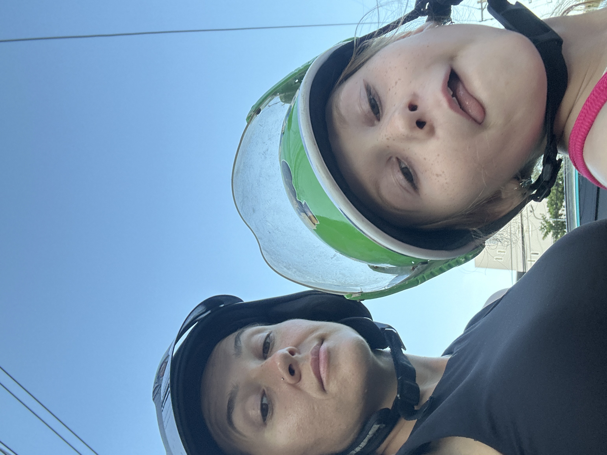 The image is a selfie taken outdoors, with a clear blue sky in the background. Two people are visible, a woman and a young girl, both wearing helmets. The woman, on the left, is looking towards the camera with a neutral expression. She has dark hair and is wearing a black tank top. The girl, on the right, is sticking her tongue out playfully. She has blonde hair with freckles and is wearing a pink top. The girl's helmet is bright green with a clear visor. In the background, partially obscured, are some buildings and overhead wires, suggesting an urban or suburban environment. The image may have been taken in the city of Bac Nha Trang Ward, Vietnam, given the presence of overhead wires and the general architectural style visible in the background.