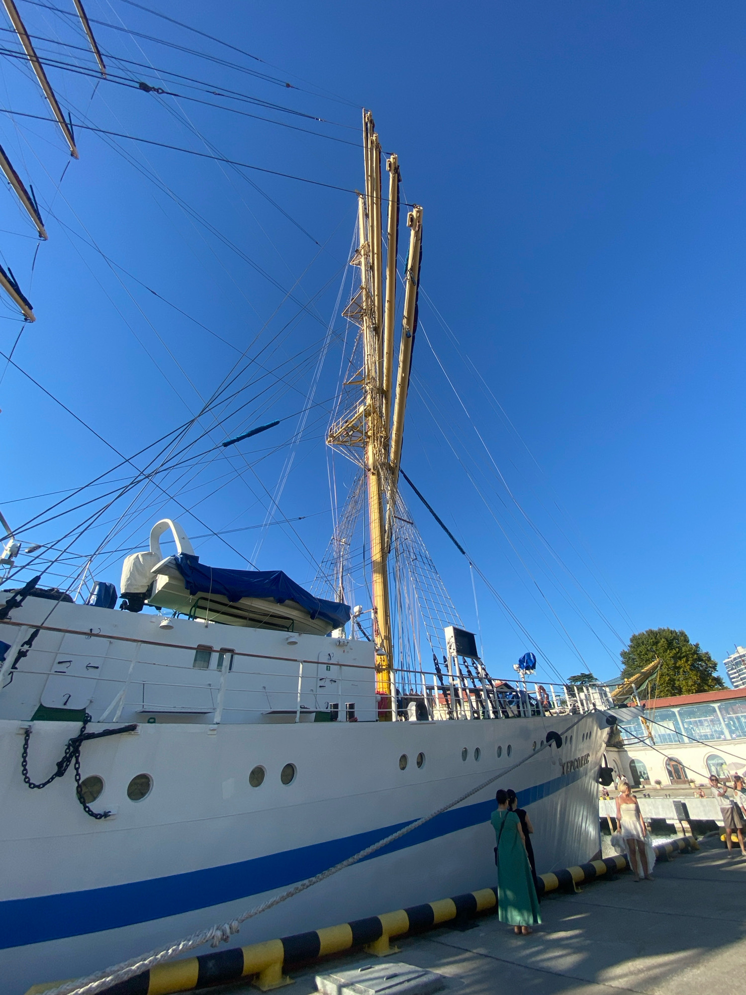 The image shows a large white ship with blue trim docked in Sochi, Russia. The ship has multiple masts and is covered in a complex network of rigging and lines. The ship's name, "Херсонес" (Khersones), is visible on the side. In the foreground, a yellow and black striped barrier runs along the edge of the dock. Several people are visible on the dock near the ship. One woman in a long green dress stands with her back to the camera. To her right, a woman in a white dress is posing for a photo. Other individuals are also present in the background, some seemingly engaged with the ship. The sky is clear and blue, and a green tree and buildings are visible behind the ship on the right side of the frame. The ship is positioned at a pier, suggesting it is either about to depart or has recently arrived. The overall scene captures a moment of activity at the port with the grand sailing ship as the central focus.