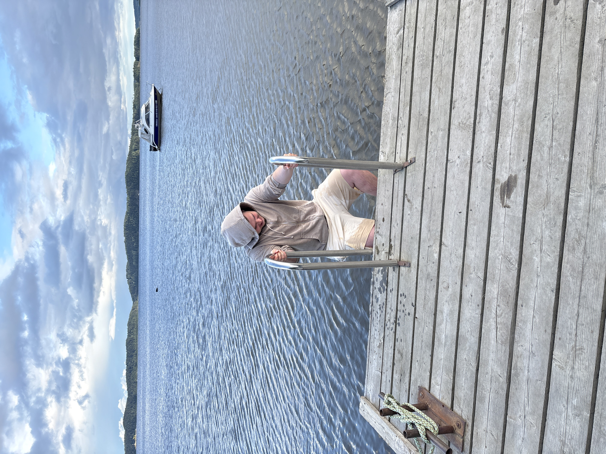 A person wearing a light brown hooded sweatshirt and light-colored shorts is partially submerged in the water of a lake, holding onto the stainless steel handles of a ladder. The ladder extends from a wooden dock into the water. The dock is constructed of weathered wooden planks. In the background, a white and blue boat with a cabin is visible on the lake. The lake is calm with gentle ripples. Distant, tree-covered hills form the horizon under a cloudy sky. The location is identified as Kir'yavalakhti, Russia.