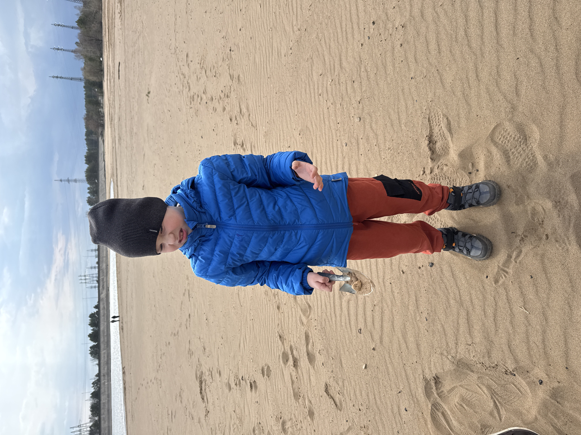 A young boy stands on a sandy beach near Noviy, Russia. He is wearing a black beanie, a blue puffer jacket, and rust-colored pants. He holds a small shovel filled with sand in his left hand. The background shows a body of water, trees, and power lines stretching into the distance. There are footprints in the sand surrounding the boy. Two people are visible walking along the waterfront in the distance.