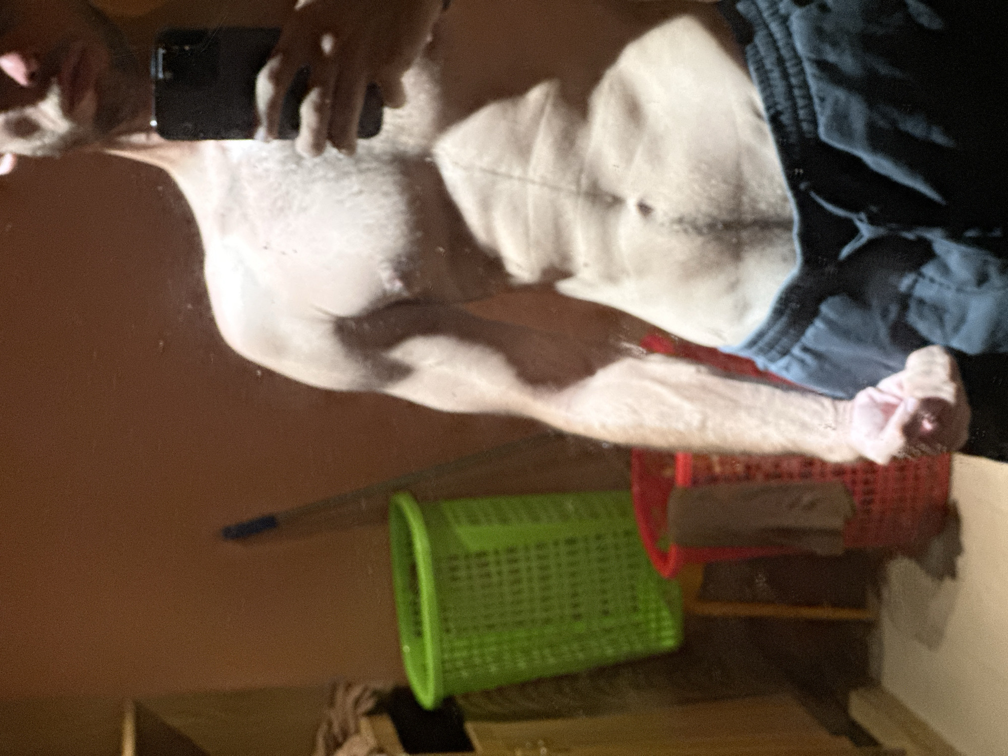 The image captures a close-up, low-angle, and horizontally oriented shot of a person's torso, reflecting in what appears to be a mirror or glossy surface. The subject, a male, is shirtless, revealing well-defined abdominal muscles and a scar across the abdomen. He is wearing dark blue shorts. In his right hand, he is holding a dark object, possibly a phone.
In the background, reflected in the same surface, are several objects. A bright green plastic basket is prominently visible, lying on its side. To its right, a red plastic basket is also visible. There are hints of furniture and other objects, suggesting an indoor setting. A long, thin, dark object, possibly a broom or mop handle, is angled across the scene. The lighting suggests an indoor environment, possibly during the day, though the exact time is not ascertainable. The reflection creates a sense of depth and perspective. No text is discernible in the image.