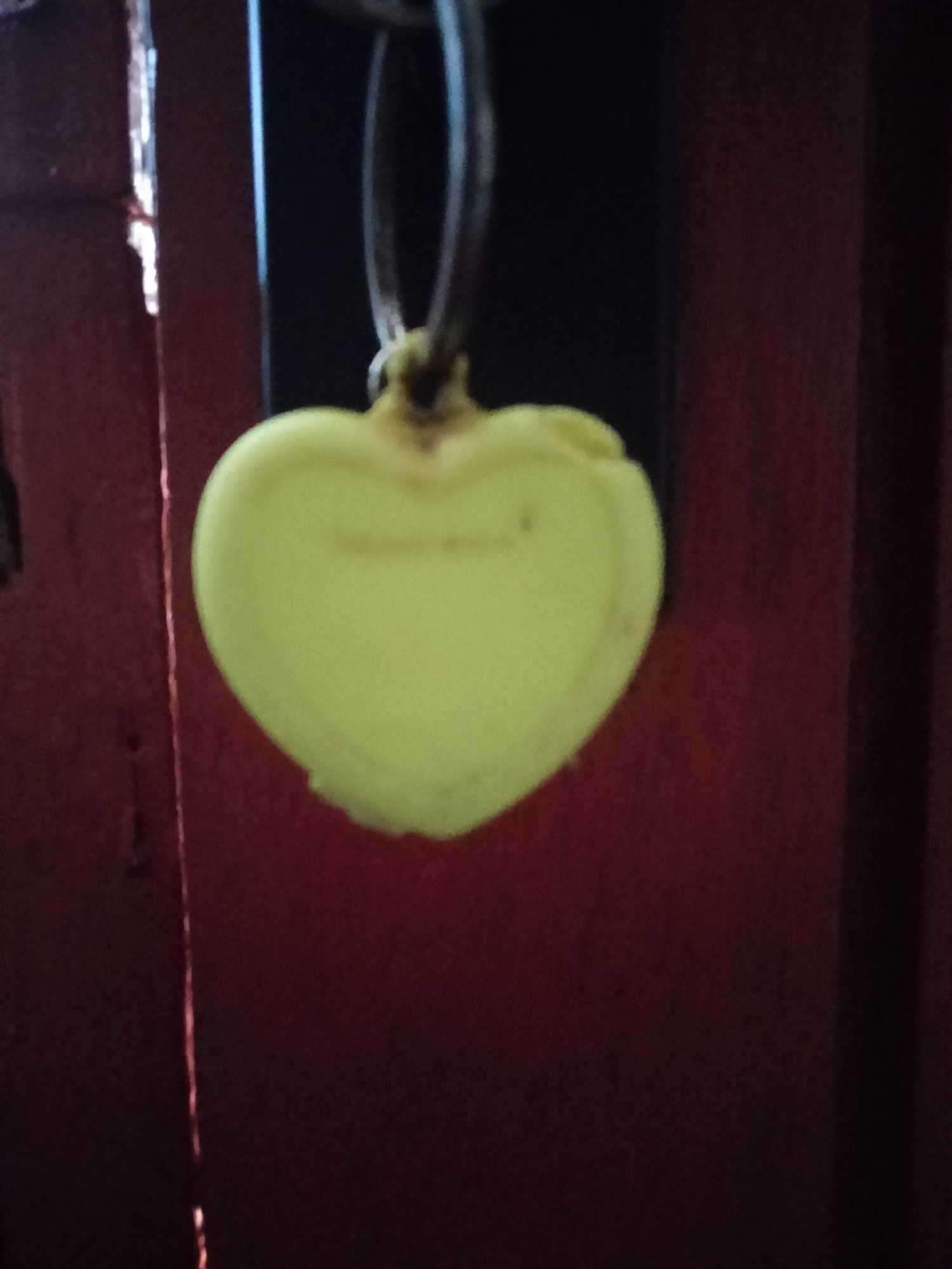 A yellow heart-shaped keychain is hanging from a metal ring against a dark red background. The background appears to be a wooden door or wall. The keychain is centrally located and slightly out of focus.