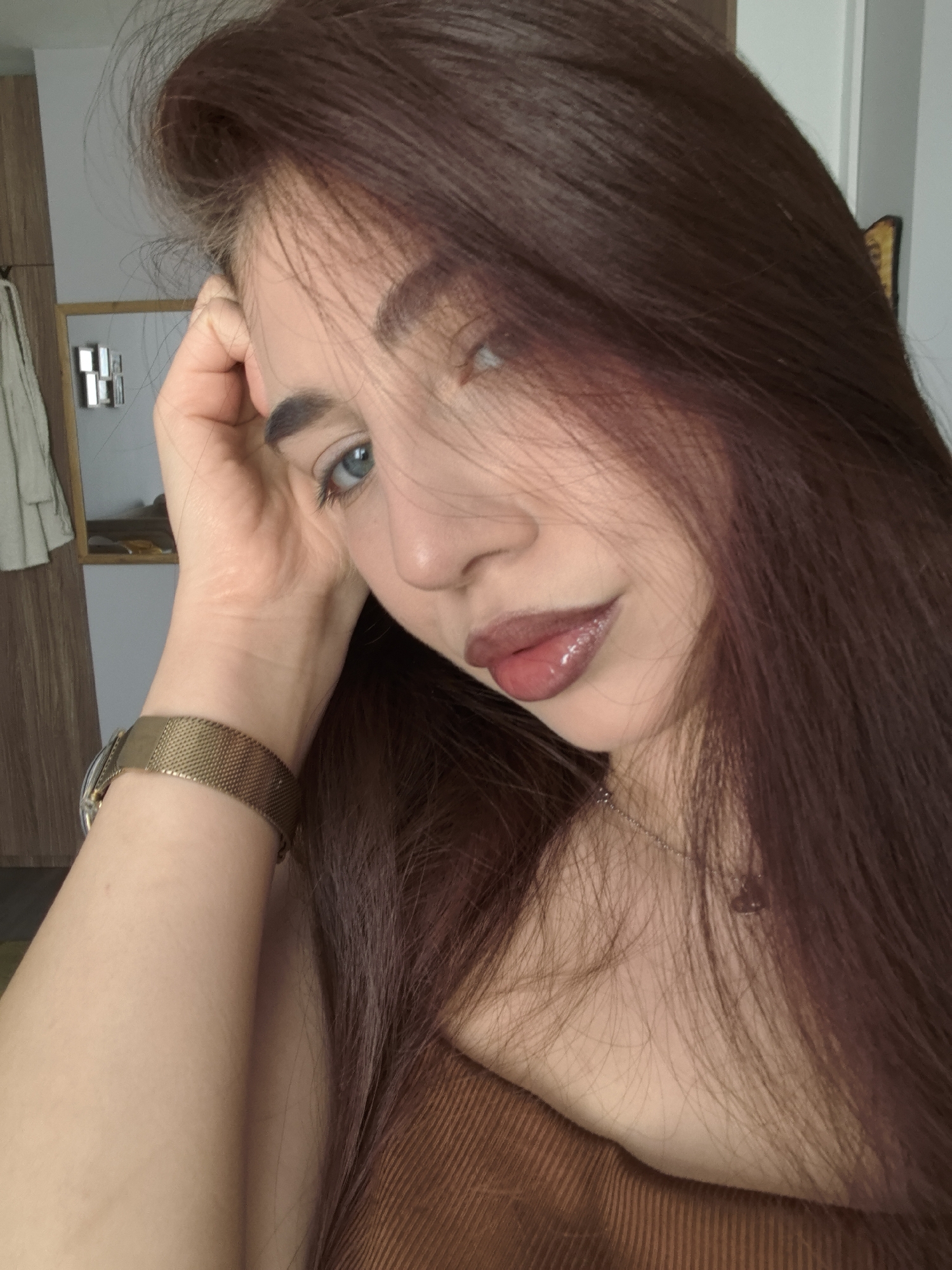 A close-up portrait features a young woman, likely a selfie, taken indoors in Stara Zagora, Bulgaria. She has long, straight, dark reddish-brown hair that cascades over her shoulders and chest, partly obscuring her body. Her fair skin is softly lit by natural light, casting subtle shadows. She possesses striking blue eyes, and her gaze is directed towards the camera with a gentle, slightly pensive expression. Her full lips are highlighted with a glossy pinkish-brown tint.
Her left hand is raised to her head, with fingers gently resting near her temple and forehead. On her left wrist, she wears a gold-toned watch featuring a finely meshed strap. A delicate silver-colored necklace with a small pendant is partially visible around her neck. She appears to be wearing a brown, ribbed top or dress.
The background is softly blurred but reveals an indoor setting. To the left, a wooden cabinet or wardrobe is visible, with a light-colored garment hanging from it. Several small, rectangular, reflective objects, possibly decorative elements or handles, are affixed to the cabinet. The scene conveys a relaxed, daytime moment, devoid of any visible text.