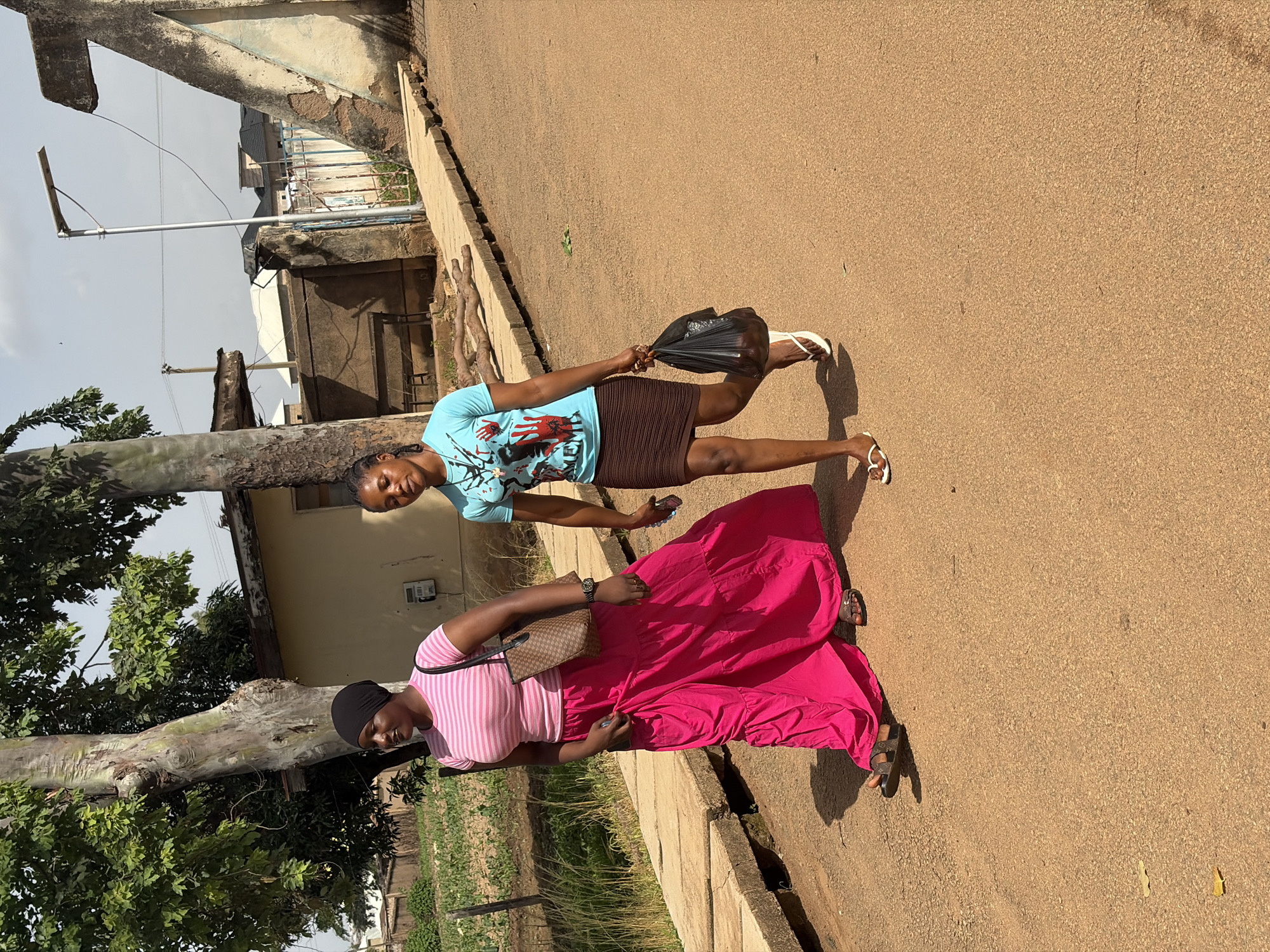 Two young women are walking on a dirt road in Rafin Par, Nigeria. The woman on the left wears a pink and white striped t-shirt and a long, bright pink skirt. She carries a brown, patterned bag over her left shoulder. The woman on the right wears a light blue t-shirt with a graphic print and a brown, ribbed skirt. She carries a black plastic bag in her right hand. Both women are wearing flip-flops. The environment appears to be a semi-urban area, with a building in the background, some trees, and a solar-powered street light visible. The ground is a mix of dirt and concrete.
