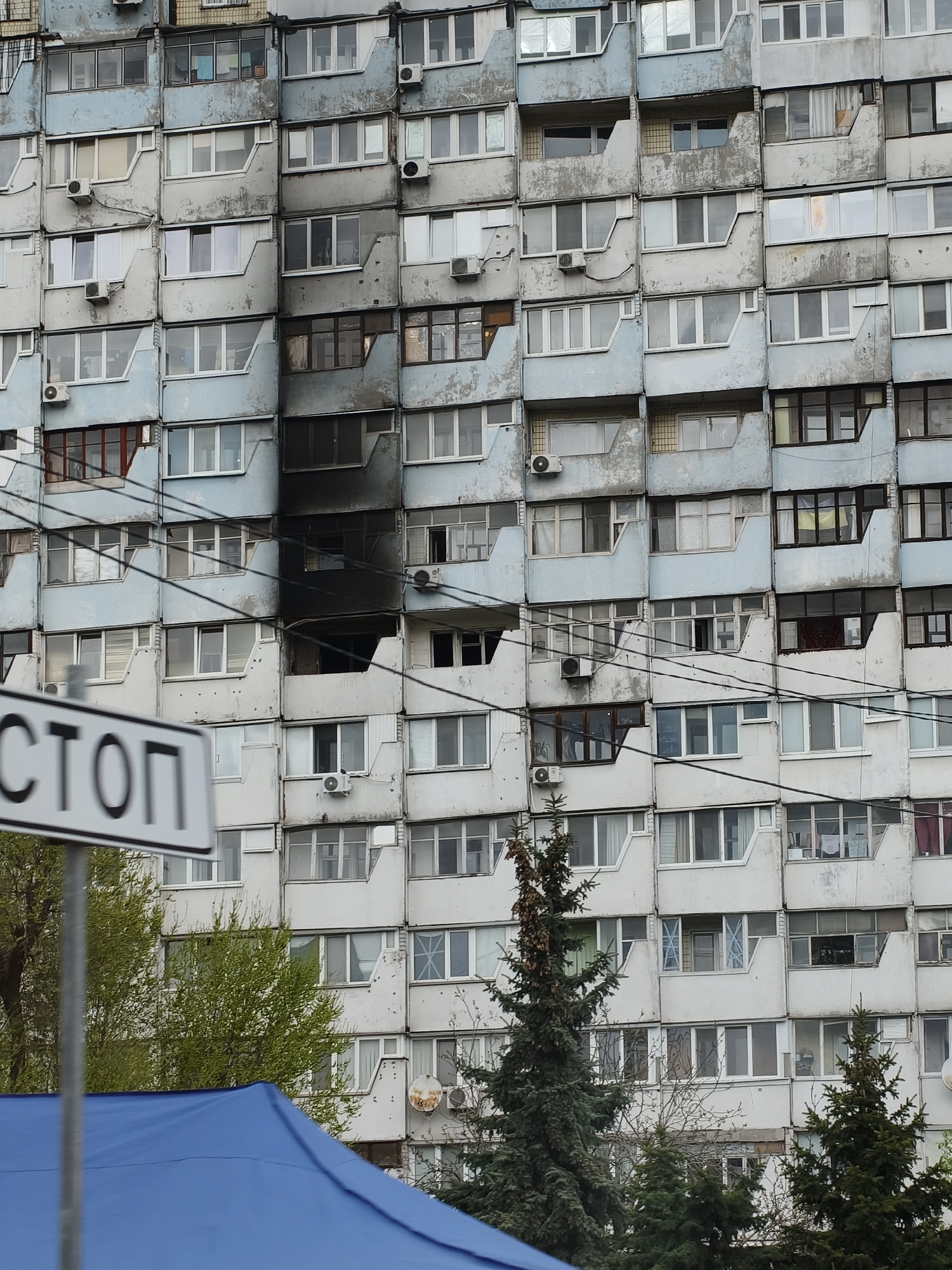 The image shows a multi-story apartment building in Dnipro, Ukraine, exhibiting damage from what appears to be a recent fire. A dark, charred vertical section runs up the facade. Windows and balconies are visibly affected, with some broken or missing. Air conditioning units are attached to the building's exterior. A "STOP" sign is partially visible in the foreground, alongside some trees and a blue tarp, suggesting the presence of emergency response or investigation activities. The overall scene conveys a sense of destruction and aftermath.