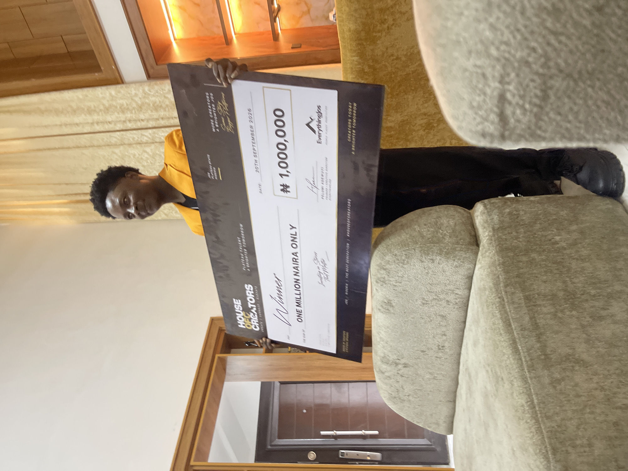 A person wearing an orange blazer and black pants stands in front of a large check and a door. The person is holding the check with both hands. The check is for one million Naira and is from "House of Creators". The person is looking to the left of the frame with a slight smile. The background shows a doorway and a wall with lighting. The scene appears to be indoors, in what looks like a modern home or office. The location of Gura Top, Nigeria, is not visible or implied in the image.