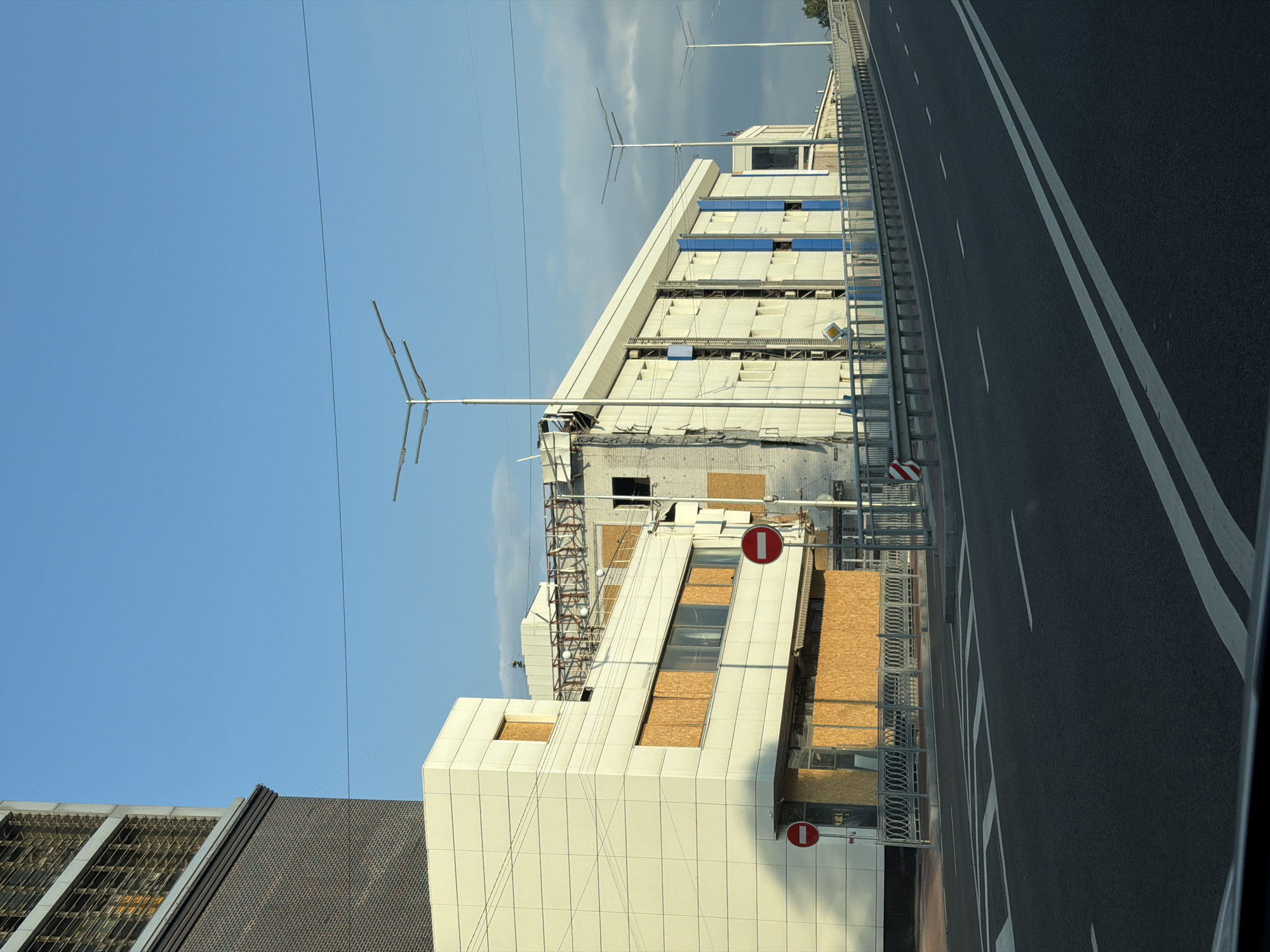 A street-level, eye-level shot captures an urban scene in Kyiv, Ukraine, under a clear blue sky with scattered clouds. The image showcases a modern building with a white facade, its windows boarded up with plywood, suggesting ongoing construction or renovation. Adjacent to it, another building, partially deconstructed, reveals exposed metal beams and brickwork, also covered with plywood.
A road in the foreground is marked with white lines, indicating lanes and pedestrian crossings. Along the side of the road, a metal railing runs parallel to the buildings. Two red-and-white "do not enter" signs are visible on posts, one to the left and another further down the street. Several streetlights with modern designs are positioned along the road. The overall impression is a city undergoing development or recovery, with elements of both new construction and visible signs of demolition or damage.