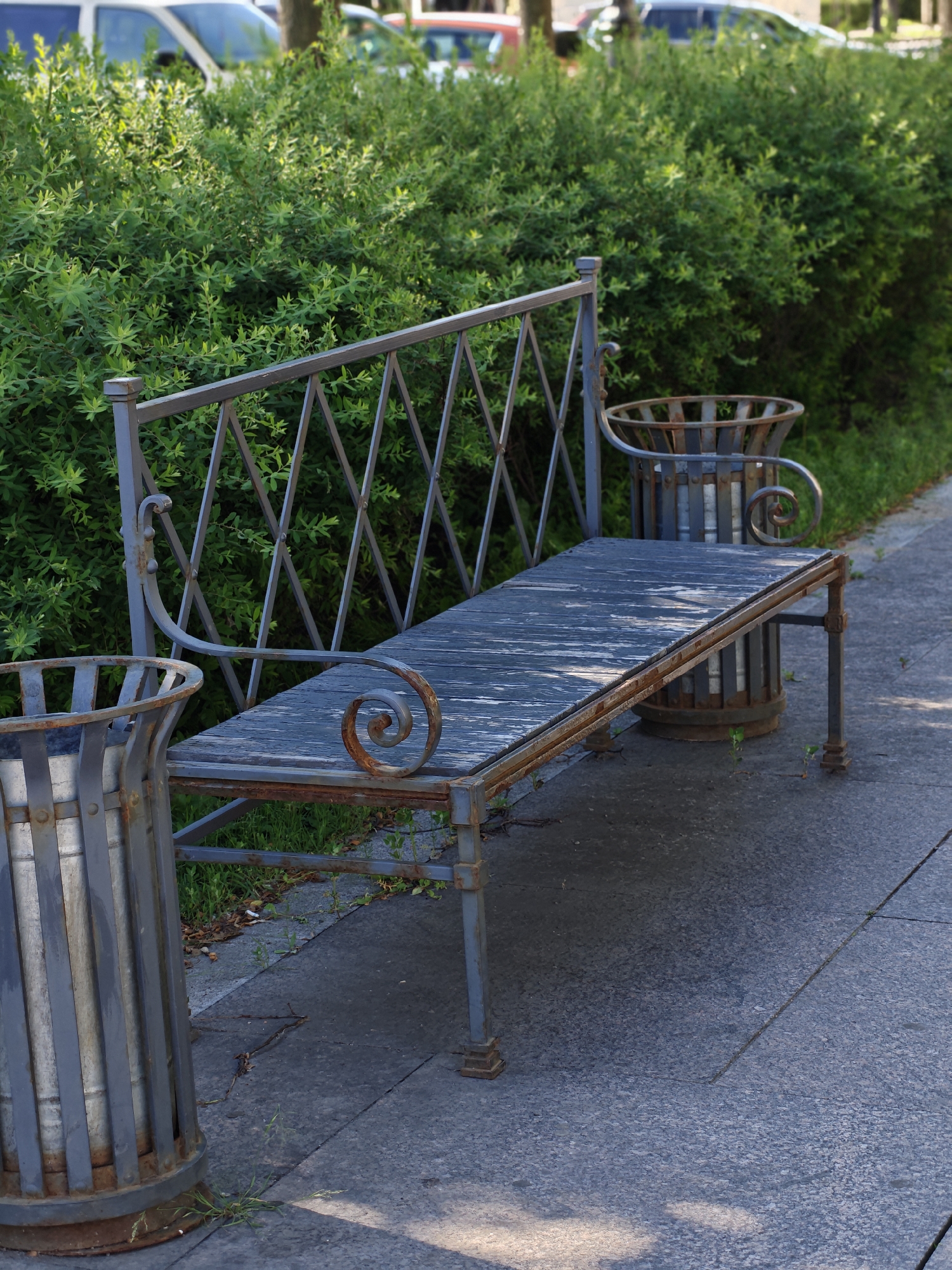 This image depicts an outdoor scene with a weathered metal park bench as the central subject. The bench has a rustic appearance with a dark blue, distressed wooden seat and a grey metal frame adorned with decorative scrollwork. Flanking the bench are two cylindrical metal trash receptacles, also showing signs of age and rust.
Behind the bench, a dense, vibrant green hedge forms a natural backdrop, creating a sense of enclosure and separation from what lies beyond. In the blurred background, hints of vehicles, possibly parked cars or vans, suggest a proximity to a road or parking area. The ground in front of the bench is paved with grey concrete or stone, with some small plants and fallen leaves visible.
The lighting suggests it is daytime, with sunlight casting shadows on the pavement, indicating a sunny day. The overall mood is tranquil and perhaps a bit melancholic due to the aged appearance of the furniture. The setting appears to be a public space, possibly a park, a sidewalk, or a garden area, in Saint Petersburg, Russia, given the context. No people are visible, and no text is present in the image.