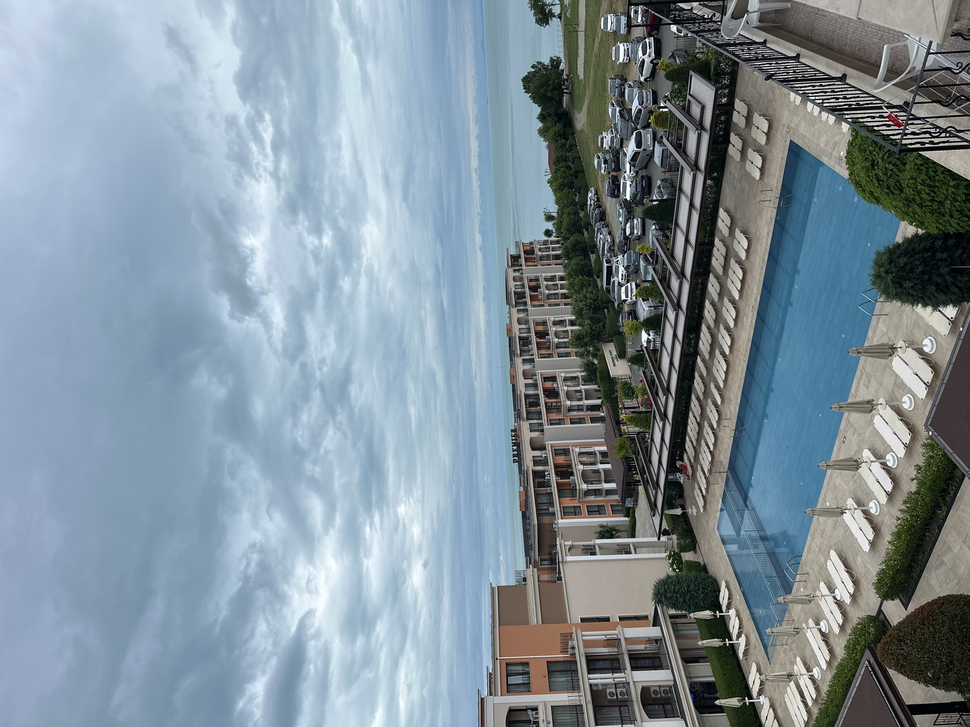 The image shows an aerial view of a resort complex in Sveti Vlas, Bulgaria, on a cloudy day. In the foreground, a large, empty swimming pool with blue water is surrounded by rows of empty lounge chairs and umbrellas. Adjacent to the pool is a parking lot filled with various cars. Behind the parking lot, a multi-story building with balconies and windows is visible, and further back, the sea stretches to the horizon under a sky filled with dramatic clouds. The architecture suggests a modern resort, with balconies on the buildings and manicured landscaping around the pool area. No people are clearly visible in the scene.