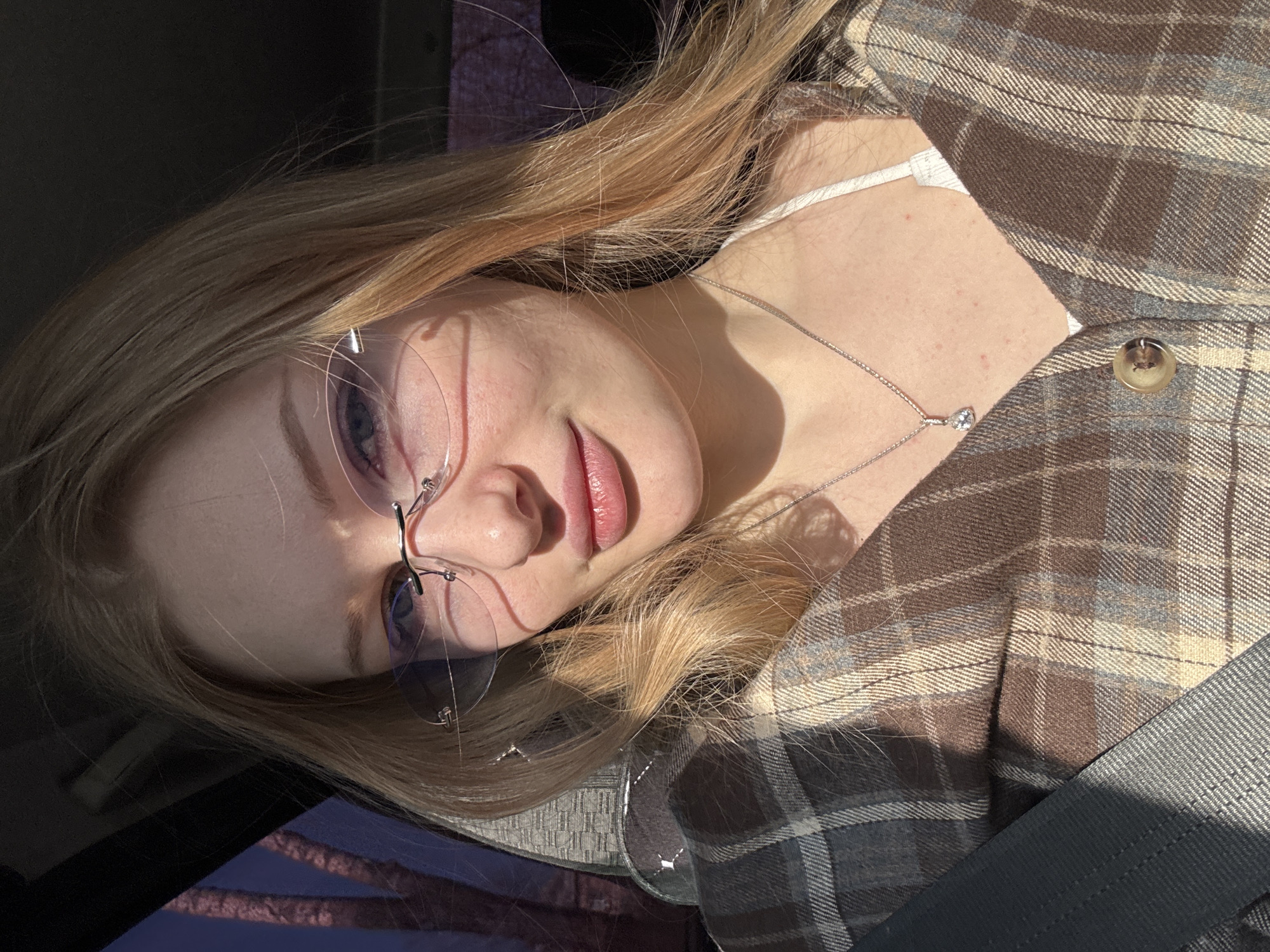 A young woman is sitting in a vehicle, wearing a brown and grey plaid blazer with a white undershirt, a silver necklace, and frameless glasses. She is looking directly at the camera with a slight smile. Sunlight is casting shadows on her face and clothing. A gray seatbelt is visible. No location information is present.