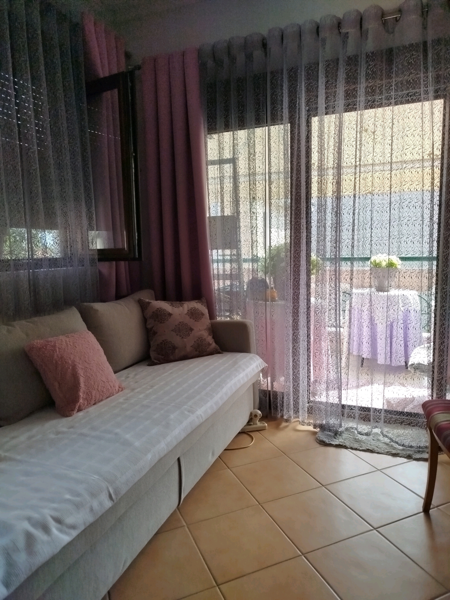 The image depicts an indoor scene, likely a living room or lounge area, with a view through sheer curtains to an outdoor patio or balcony. The foreground features a light-colored sofa with several cushions. One cushion on the left is a muted pink, while another on the right is patterned with a darker motif. The sofa is covered with a white striped throw. Adjacent to the sofa is a tiled floor in earthy tones.
Through the sheer, textured curtains, an outdoor area is visible. There are two tables, one covered with a light purple tablecloth, and a white pot with a floral arrangement on it. The overall impression is of a relaxed and somewhat decorated space. The location is identified as Sithonia Municipal Unit, Greece.