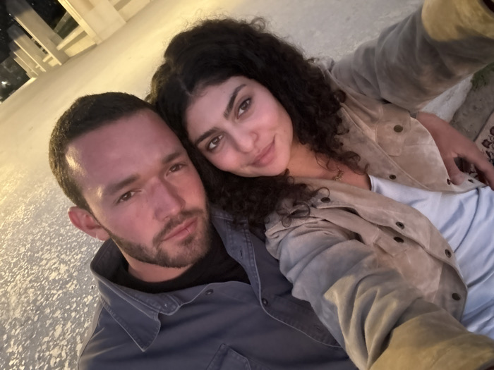 A selfie-style photograph depicts one adult male and one adult female positioned closely in the foreground against a backdrop of illuminated architectural structures. The scene is identified as the Getty Center in Los Angeles, United States of America, evidenced by the characteristic white stone balustrades and buildings visible under warm artificial lighting at night.
The male subject, positioned on the left, has short dark hair, a stubble beard, and is wearing a dark gray collared shirt. He looks directly at the camera with a neutral expression.
The female subject, positioned to the right and slightly behind the male, has long, dark, curly hair. She is wearing a light beige or tan suede-like jacket over a light-colored top, with a necklace visible. She smiles softly and looks directly at the camera, leaning her head towards the male subject.
The immediate ground surface is light-colored and textured, possibly stone or concrete. The overall lighting suggests an evening or nighttime setting, with artificial light sources illuminating the subjects and the architectural elements.