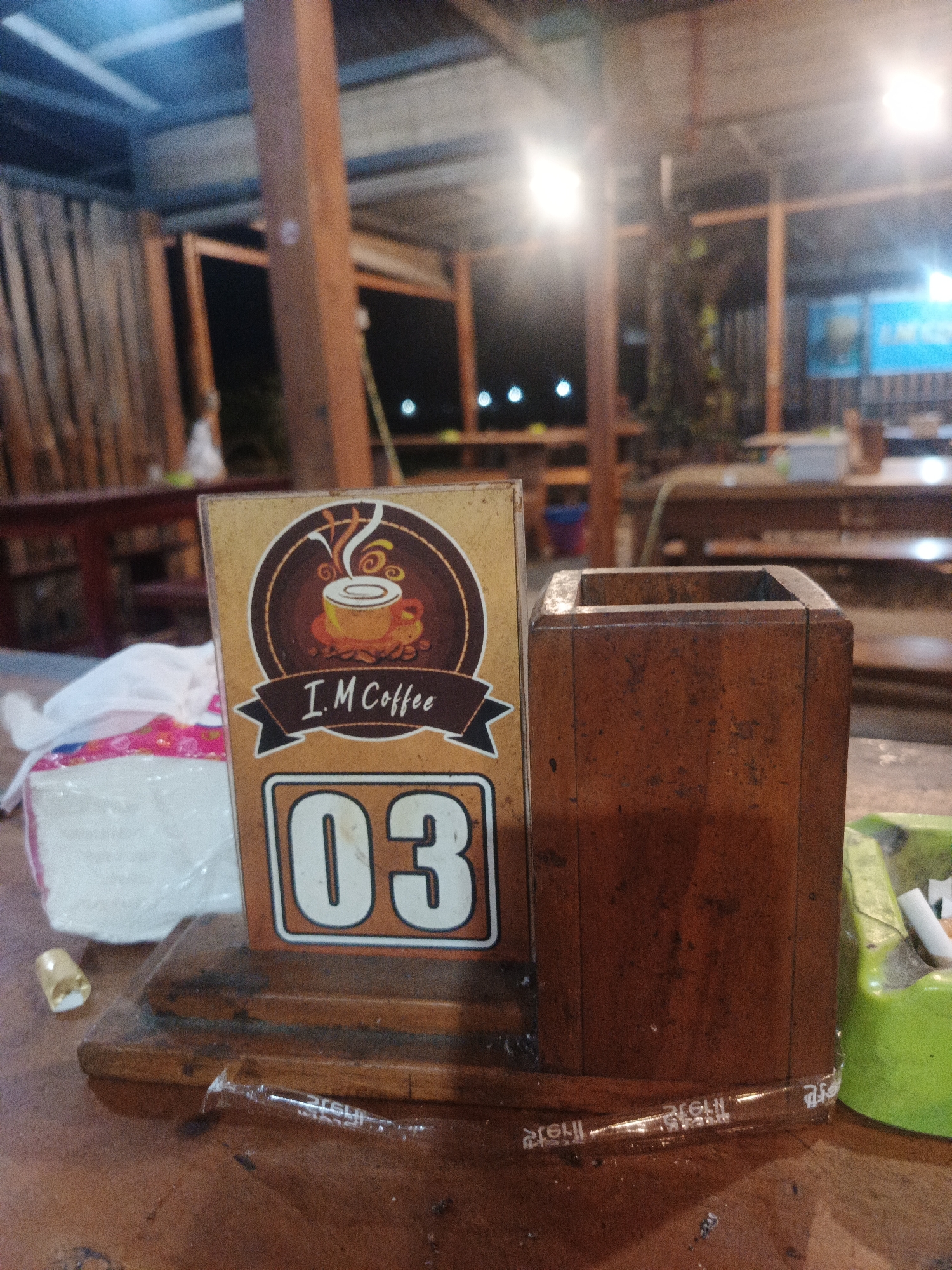 The image presents a close-up view of a wooden table at what appears to be a rustic outdoor or semi-outdoor cafe or coffee shop, likely at night given the artificial lighting. The scene is set in Geulumpangmeujim-Jim, Indonesia.
In the foreground, a brown table stand is prominently displayed, featuring a logo with a stylized coffee cup and steam, surrounded by a dark brown circle. Below the logo, a banner reads "I.M Coffee," and beneath that, the large numbers "03" are visible. To the right of this stand, a simple, rectangular wooden container or holder sits on the table.
On the left side of the table, a pack of white tissues, wrapped in plastic with a hint of pink pattern, is present. A small, light-colored crumpled object lies near the tissue pack. To the far right, a bright green ashtray containing several cigarette butts is partially visible. A transparent plastic wrapper, possibly from a straw, lies flat on the wooden table surface in front of the stand, showing some mirrored, indistinct text that appears to be a brand or product name (e.g., "IRELU").
The background is softly blurred, revealing a structure constructed from wooden beams and supports. Multiple bright artificial lights illuminate the space, casting a warm glow and creating bokeh effects. Faint outlines of other tables and chairs can be discerned in the background, suggesting a casual dining or gathering area. No people are visible in the image.