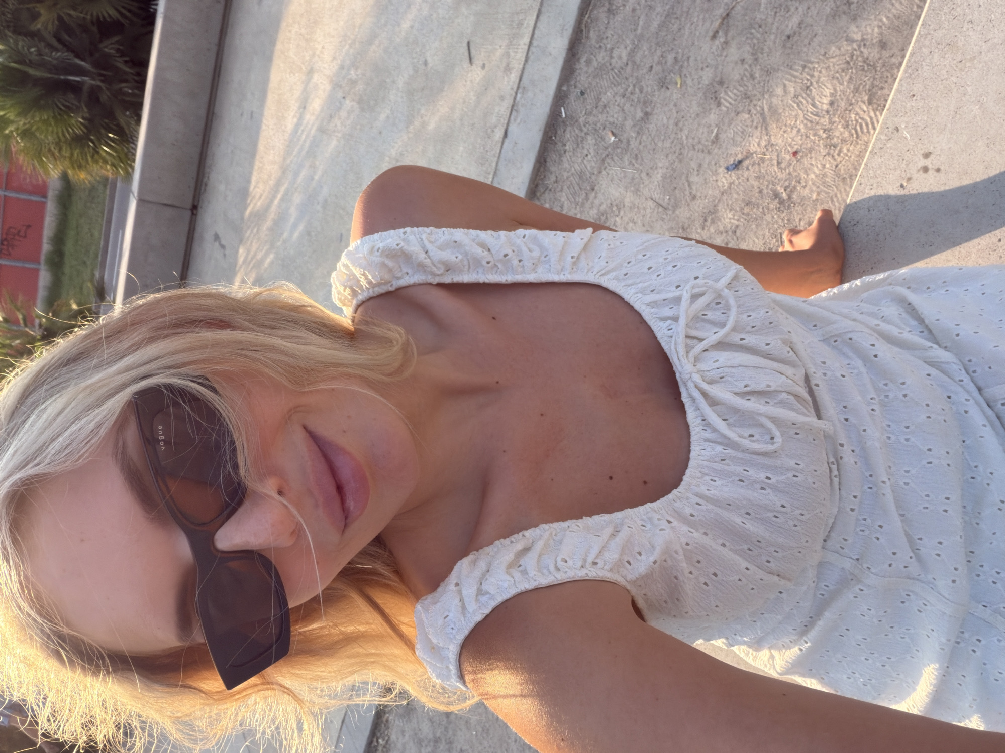 The media file is a selfie of a young woman with blonde hair and sunglasses. She is wearing a white, eyelet fabric dress with a ruffled neckline. The woman is outdoors, seated on concrete steps or a bench. The lighting suggests it is late afternoon or early evening, with warm sunlight casting shadows. In the background, there are hints of greenery, possibly palm trees, and a red brick wall with graffiti. The overall scene appears to be a casual, sunny day in an urban or park-like setting. The woman's expression is relaxed and she is looking towards the camera. There is visible text on the side of her sunglasses, which reads "VOGUE". The location context is identified as Benimàmet-beniferri, Spain.