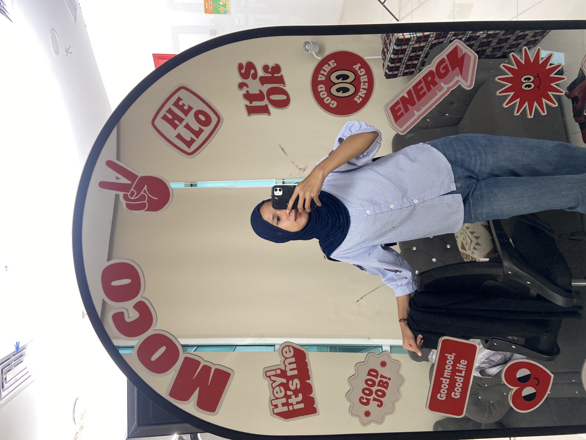 A woman wearing a blue hijab and a striped button-down shirt is taking a selfie in a mirror. She is holding her phone up to her face with her right hand, and her left hand is partially visible at the bottom right of the frame. She is wearing blue jeans. The mirror is decorated with various red and white stickers with messages like "Hello", "It's Ok", "Good Vibe Energy", "Energy", "Moco", "Hey! It's me", "Good Job!", and "Good mood, Good Life". The reflection in the mirror shows a checkered fabric, a grey couch, and a black jacket or cloth draped over a chair. The mirror is located in North Jakarta, Indonesia, according to the metadata.