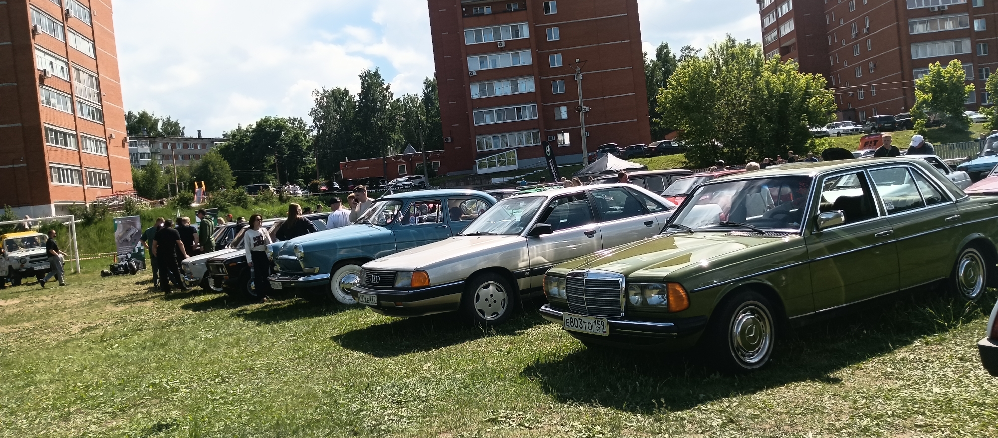 A long shot shows a group of vintage cars parked on a grassy field in a city in Unknown, Unknown, with several people milling around the vehicles. The cars are arranged in a row, and include a yellow jeep-like vehicle, two dark sedans, a blue car with white wall tires, a silver sedan, and a green sedan. The license plate of the green sedan, located at the right edge of the image, reads "E803TO 159" in Cyrillic characters. Buildings, including tall, brick apartment blocks, are visible in the background. The weather appears to be sunny.