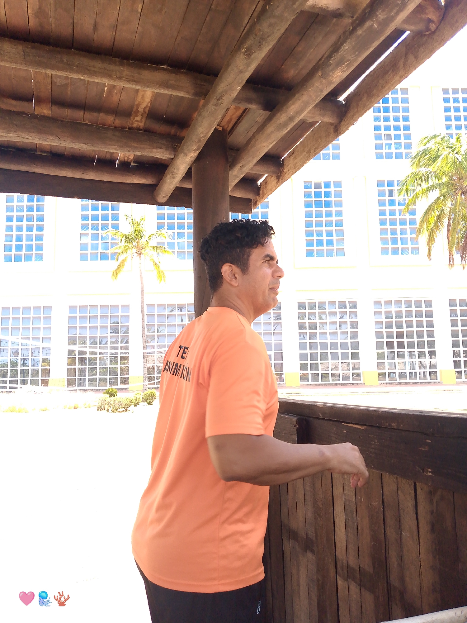 A medium shot depicts a man in an orange t-shirt standing under a wooden structure, possibly a gazebo or a covered area. He is facing to the right, gazing outward, with his left hand resting on a wooden railing. The building in the background is a large, light-colored structure with many windows, suggesting an institutional or commercial building. A palm tree is visible on the right side of the frame. The sky is bright, indicating daylight. The location appears to be Pedro González, Venezuela, though that information is not evident in the image itself.