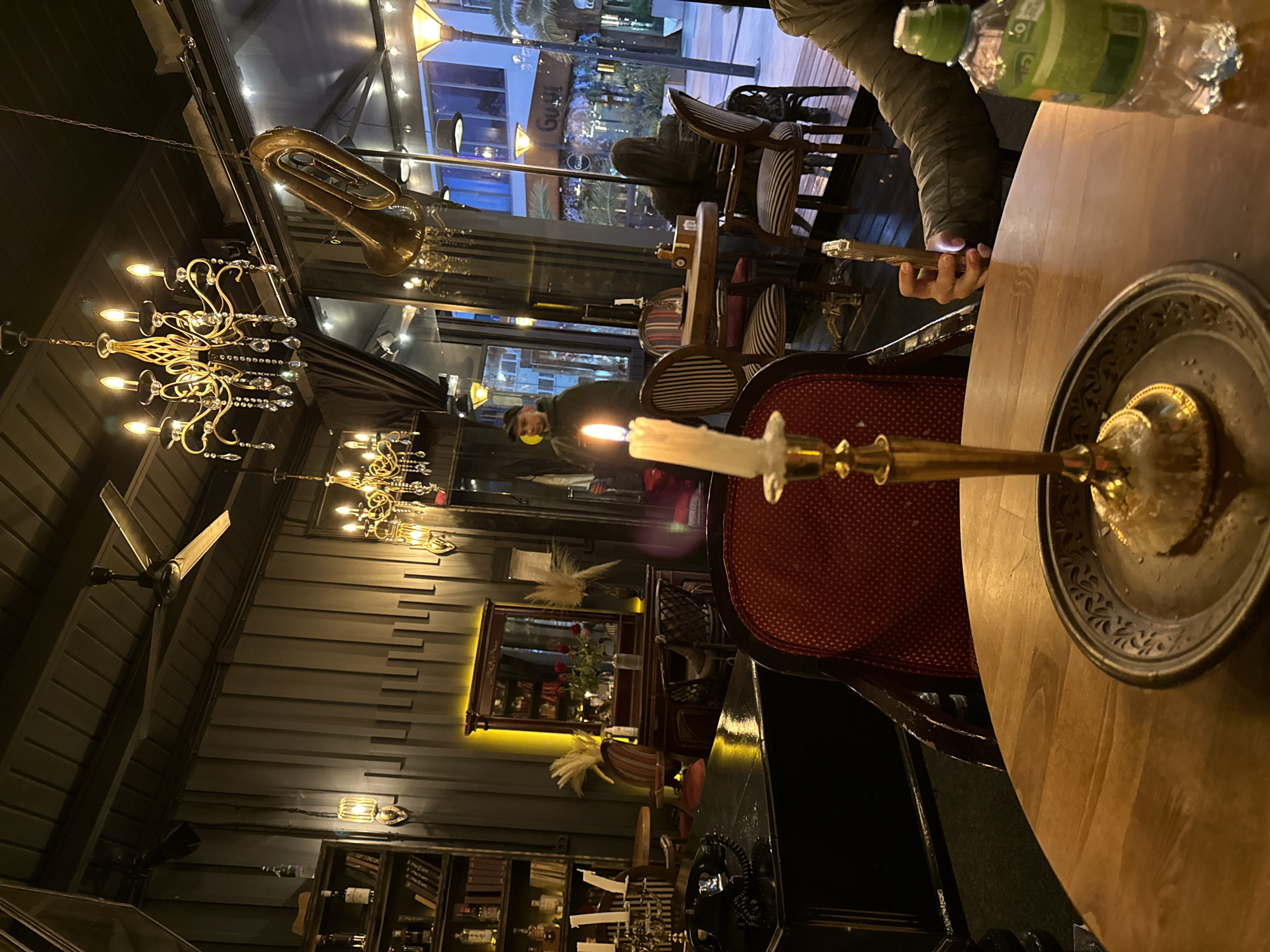 A person at a round wooden table in what appears to be a restaurant. A burning candle in a gold holder is on the table along with a water bottle and a small plate. The person is holding a phone. There are red patterned chairs. In the background are bookshelves and a mirror. Chandeliers and other decorative items, including a trumpet, adorn the ceiling. Other people are visible in the background, and the outside cityscape is visible through the windows. This appears to be a scene from Sochi, Russia.