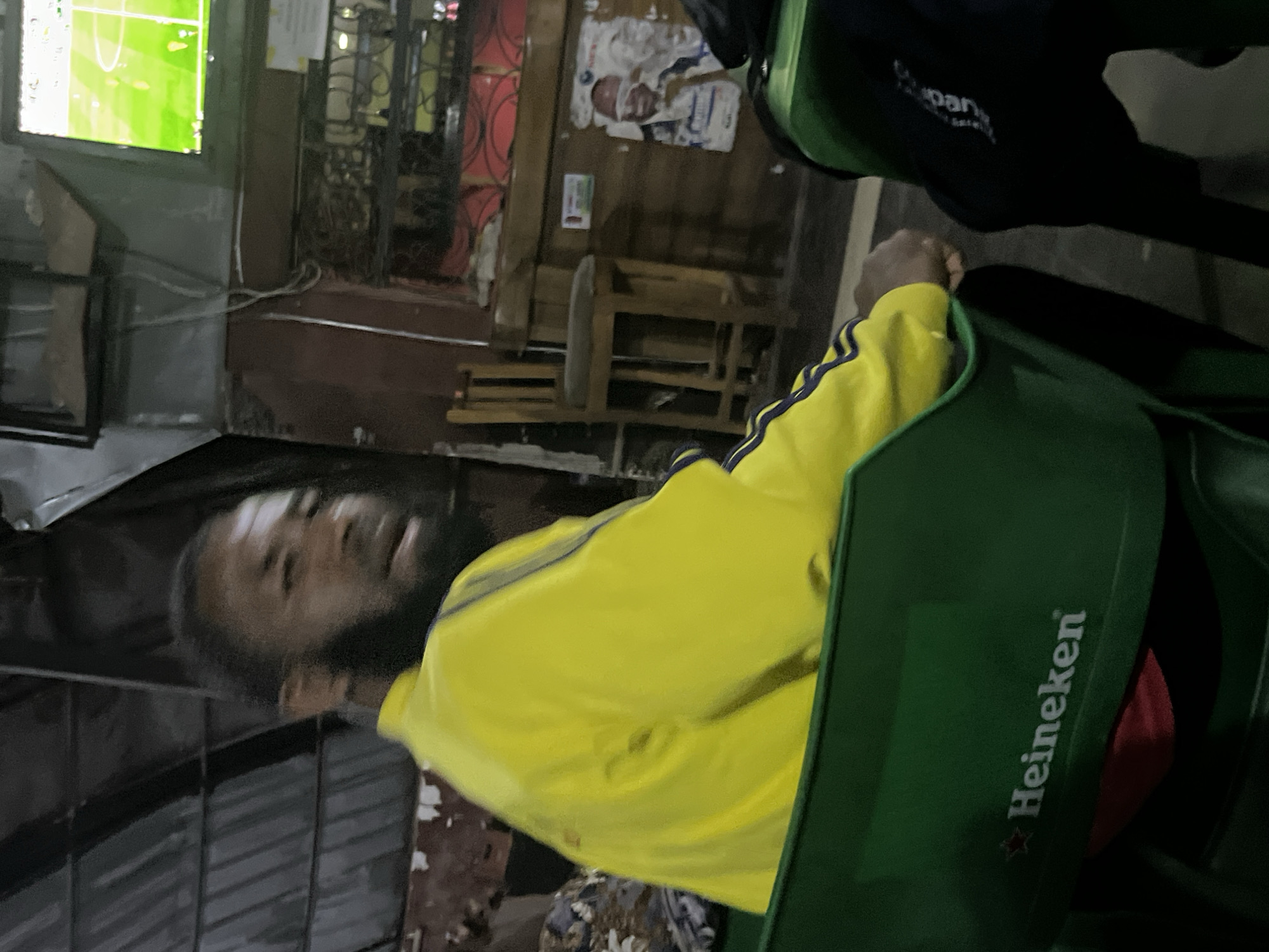 The image depicts a man wearing a yellow jacket with blue stripes on the sleeves, seated in a green Heineken chair. He has a beard and is looking towards the right. Behind him, a television screen displays a soccer match. The environment appears to be an indoor seating area, possibly a bar or a viewing center, with a wooden bar counter, chairs, and a corrugated metal roof. A poster is visible on the wall behind the bar. The scene suggests a casual setting where people gather to watch sports. The location is identified as Gura Top, Nigeria.