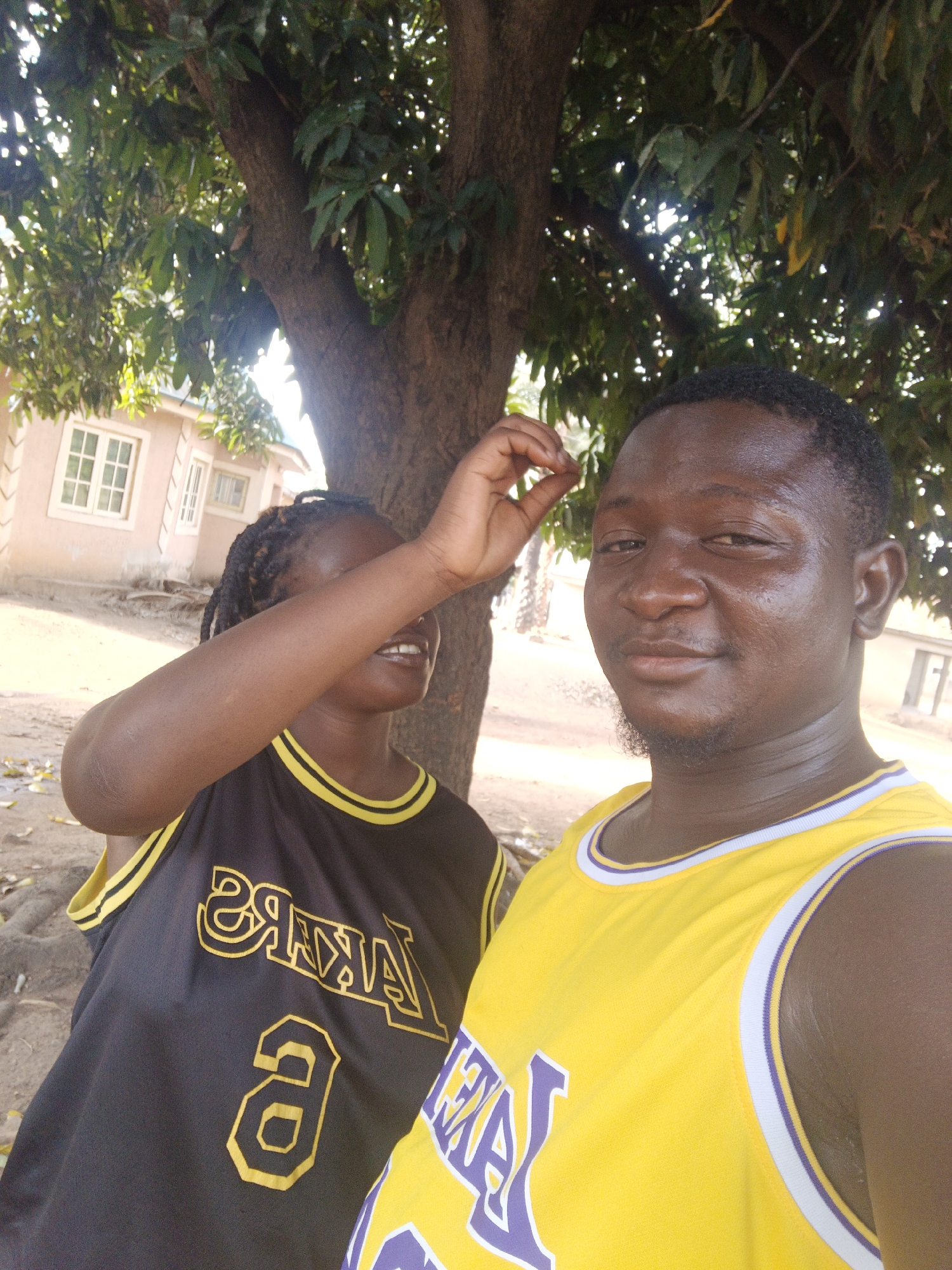Two people are posing for a photo outdoors in Akwanga, Nigeria. The individual on the right is male, wearing a yellow Lakers jersey, and is facing the camera with a neutral expression. The person on the left, possibly female, wears a black Lakers jersey and has their arm raised, covering their eyes. They are standing close to a tree with dense foliage, with buildings visible in the background. The overall scene is bright and appears to be taken during daylight. The location is a dusty area in Akwanga, Nigeria.