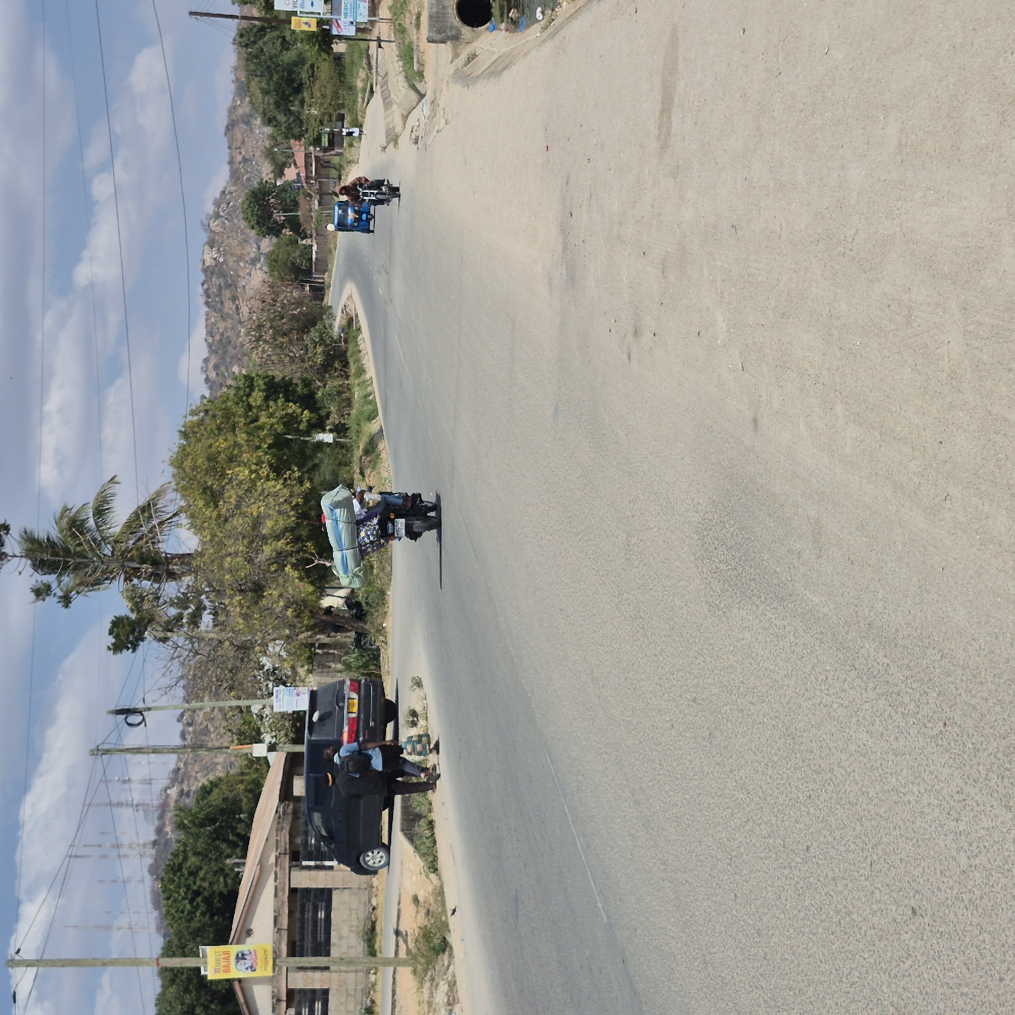 The image depicts a semi-arid urban environment in Dodoma, Tanzania. A paved road curves through the scene, flanked by sparse vegetation, residential structures, and utility poles with numerous antennas visible on a distant hill.
In the foreground, two individuals walk along the side of the road, near a dark-colored minivan. Further down the road, a person is riding a motorcycle loaded with large, bundled items, including what appears to be a mattress or sleeping bag. A blue auto-rickshaw is also visible on the road, with a person driving it.
The sky is partly cloudy, with fluffy white clouds against a pale blue backdrop. The overall impression is one of a typical day in a developing urban area, with a mix of modern vehicles and modes of transport.