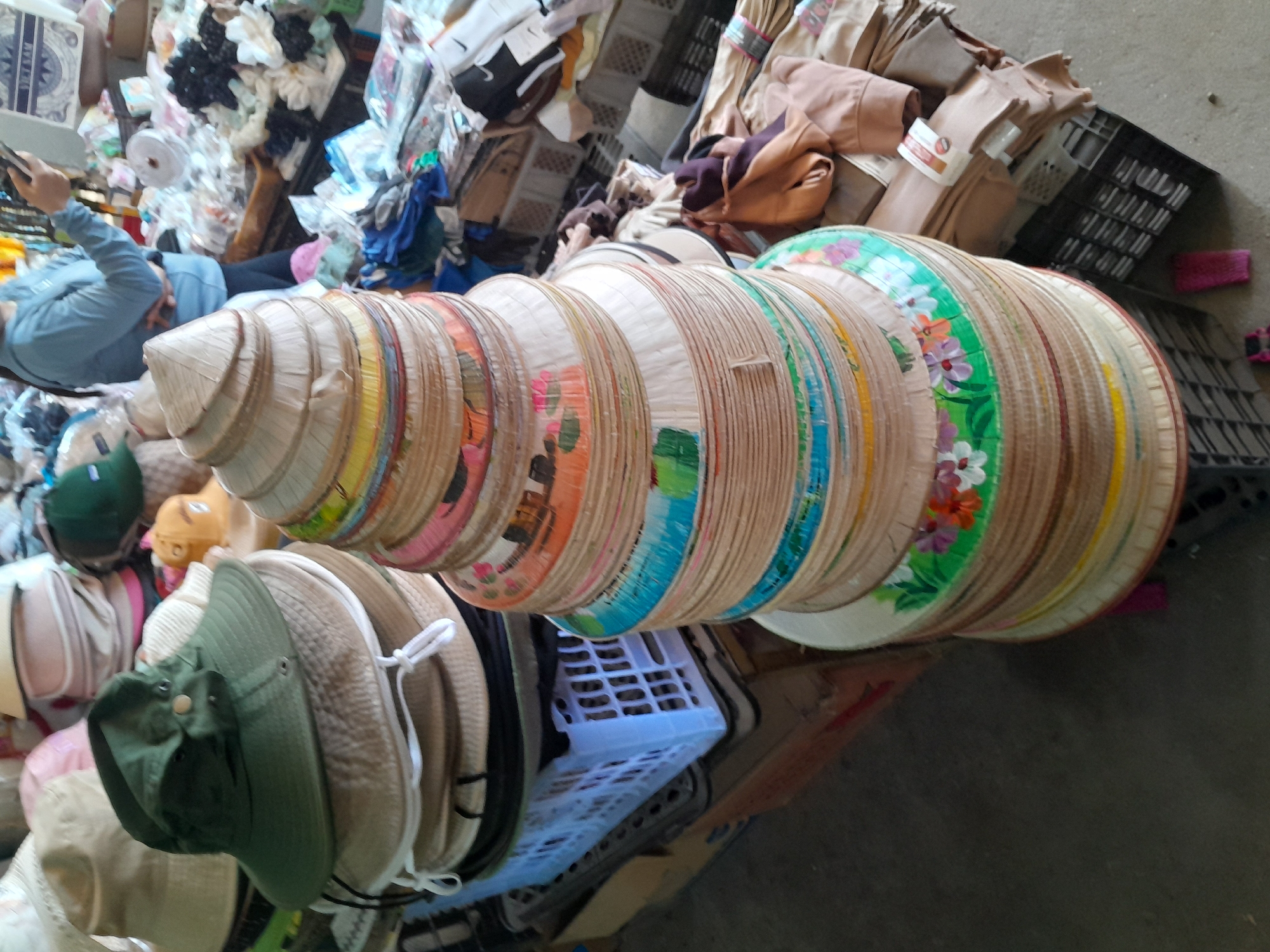 In Phu Thuy Ward, Vietnam, a market stall displays a large stack of conical hats. The hats, made of woven straw, vary in size and are decorated with colorful painted designs of flowers and leaves. A green bucket hat sits atop a smaller stack of hats to the left, and other hats and fabric items are visible in the background, suggesting a retail environment. A person in a blue shirt is partially visible in the upper right corner of the image, holding a mobile phone.