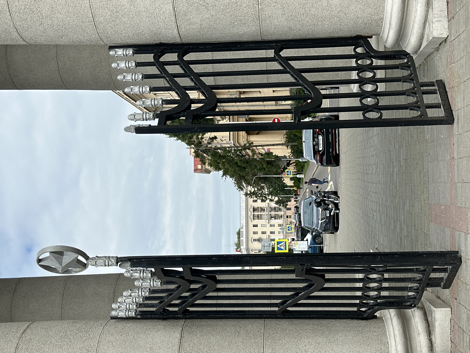 The image shows an open ornate black metal gate leading to a street in Minsk, Belarus. The gate is flanked by two large, light-grey, cylindrical columns. Atop the left side of the gate, a silver five-pointed star emblem is visible against a cloudy sky. The gate itself features vertical bars and decorative elements, including a circular motif towards the bottom.
Beyond the gate, a paved street with a brick pattern is visible, lined with parked cars and a motorcycle. Pedestrian crosswalk signs are present on the street. Several buildings are visible in the background; one prominent building on the right appears to be a light-yellow, multi-story structure with arched windows. Trees and streetlights also line the street.
In the mid-ground, a person in dark clothing is walking away from the camera on the right side of the street. A black car and a motorcycle are parked in the middle of the street. Another black car is parked on the left side of the street. The sky is overcast, suggesting a diffused light condition.