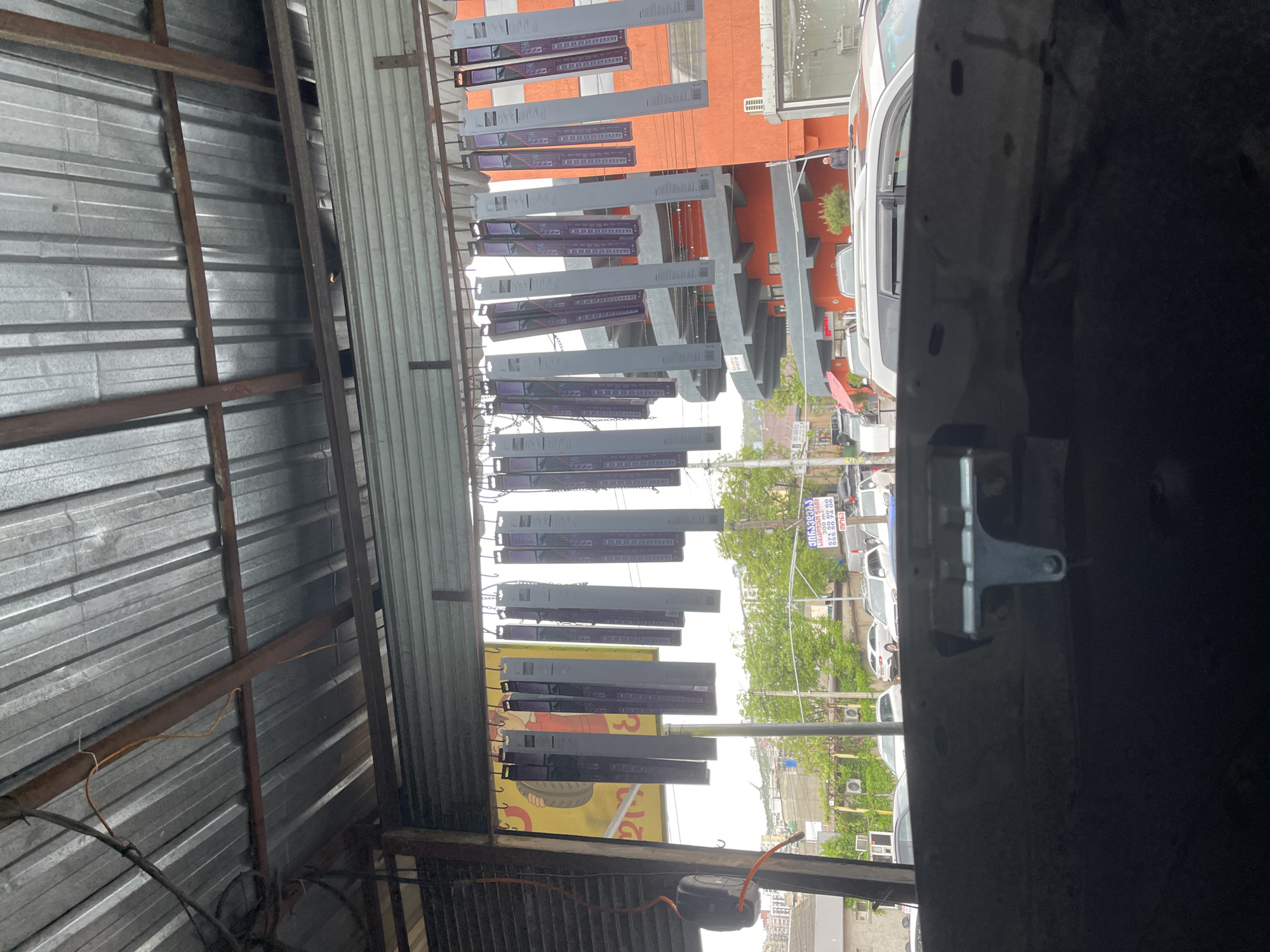 The image is taken from inside a structure with a corrugated metal roof and metal supports, looking outwards towards a street scene in Tbilisi, Georgia. Hanging from the ceiling of the structure are multiple gray rectangular objects, possibly packaged goods, suspended by chains. In the background, an orange building with gray structural elements is visible, along with cars parked on the street and green trees lining the sidewalk. The photograph is taken from a low vantage point. Part of a vehicle is obscuring the right side of the frame.