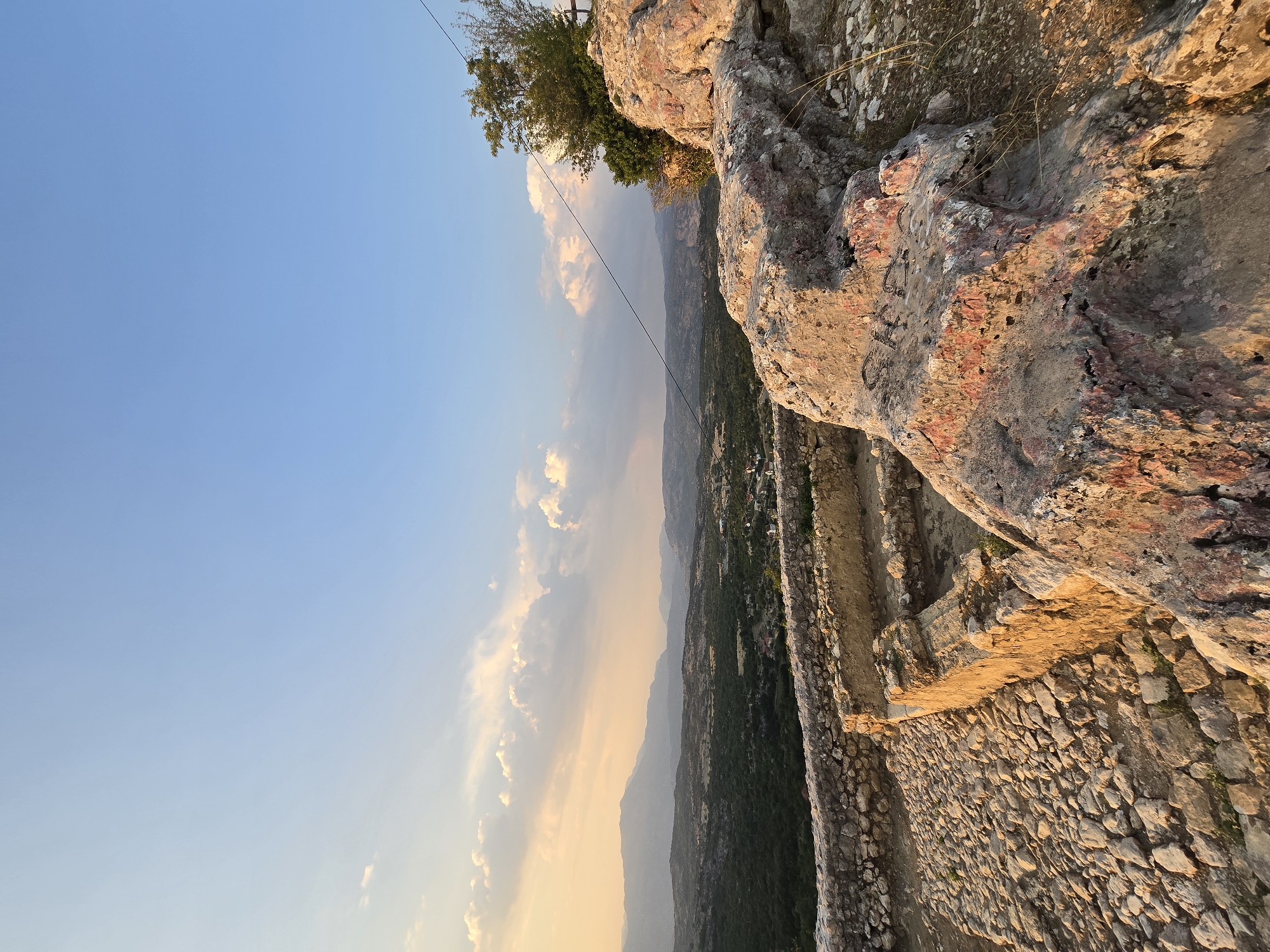 The image depicts a scenic view from an elevated position overlooking a valley. In the foreground, there are ancient stone ruins, characterized by rough-hewn stone walls and a cobblestone pathway. Large, weathered rocks with reddish and greyish hues are prominent, with some showing signs of discoloration and rough textures. A tree with green foliage grows on the right side, partially obscuring a thin wire that extends across the frame.

The middle ground features a valley with lush green vegetation, interspersed with scattered houses and buildings, suggesting a small settlement. Rolling hills and mountains form the backdrop, fading into the distance under a clear blue sky. The sky is partly filled with soft, wispy clouds, illuminated by the warm, golden light of either sunrise or sunset. The overall scene conveys a sense of historical depth and natural beauty. The location is identified as Gornja Vrbica, Montenegro.