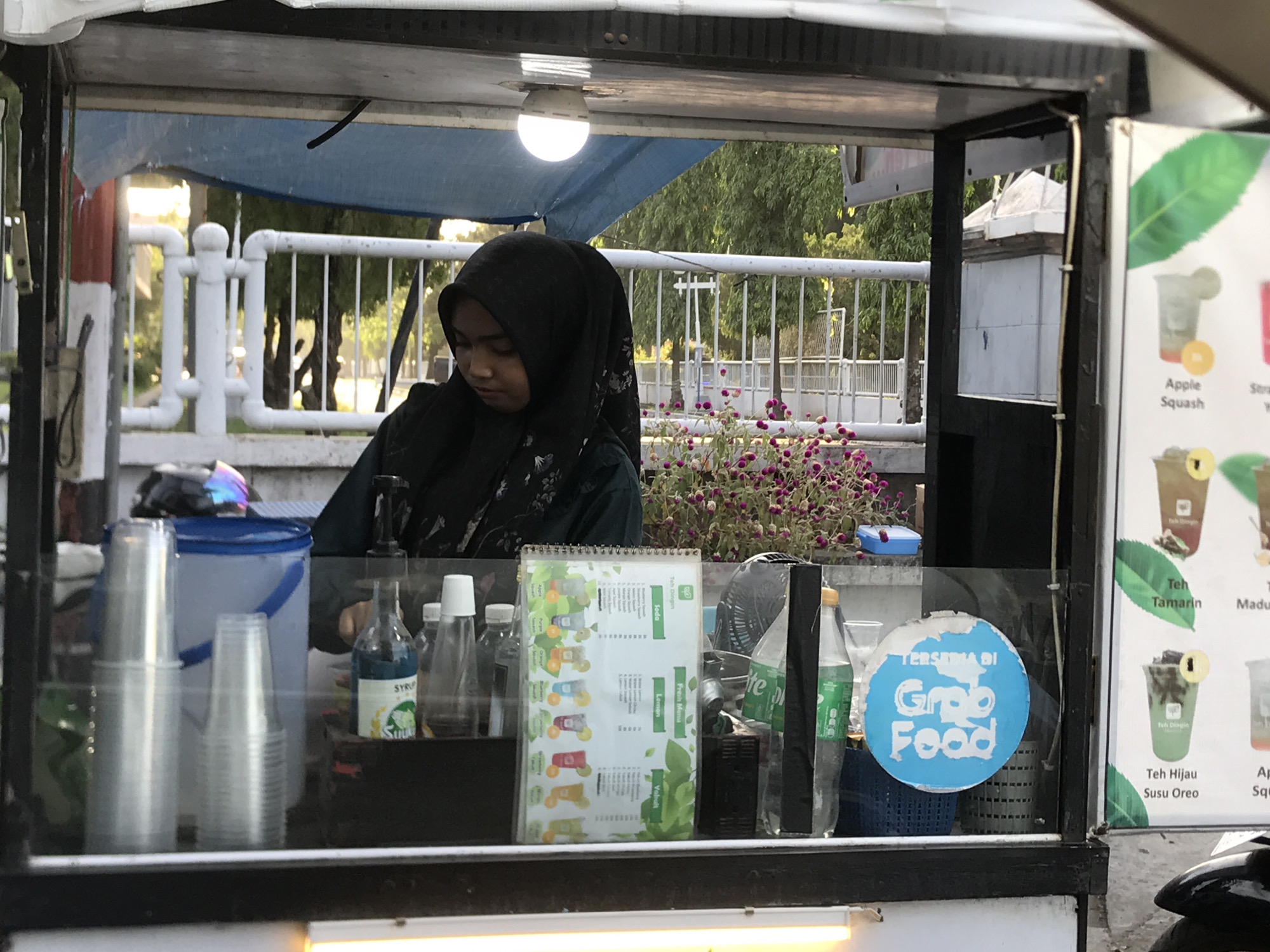 The image shows a food vendor in Lhokseumawe, Indonesia. A woman wearing a black hijab stands inside the food stall, behind a glass barrier displaying cups, syrup bottles, a drink menu, and a "Grab Food" sign. The stall is illuminated by a light. Outside the stall, there is a white fence, some trees, and purple flowers in a flowerbed. A scooter is parked on the right side of the stall.