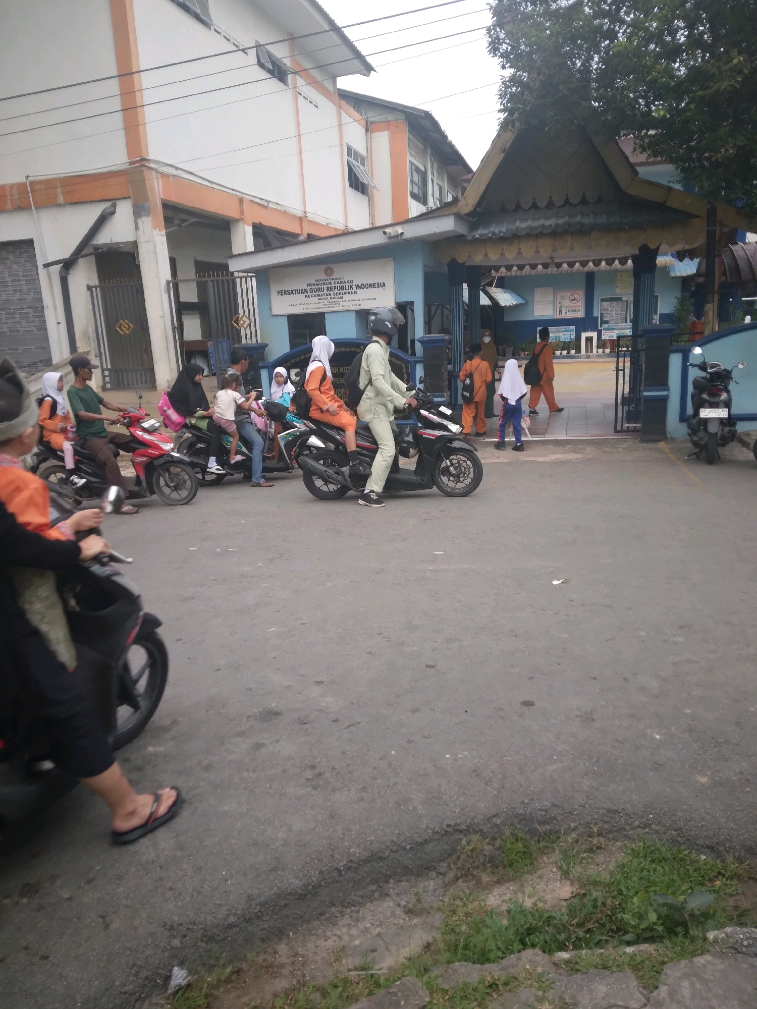 The image depicts a street scene in front of a building with an orange and white facade, likely an educational institution or government office, in Batam, Indonesia. A sign above an entrance reads "SEKRETARIAT PERSATUAN GURU REPUBLIK INDONESIA" (Secretariat of the Indonesian Teachers Association), with an address and contact information. Several people are present, most of whom are on or around motorcycles. Some individuals are wearing school uniforms, predominantly orange and white.
In the foreground, a person on a black motorcycle is partially visible from the waist down, wearing black pants and flip-flops. Several other motorcycles are in the mid-ground, some carrying multiple passengers. A person in light green clothing is riding a black motorcycle, wearing a helmet. Another motorcycle, red, carries a man in a green shirt and a woman in an orange outfit. A woman in a black headscarf is riding on the back of a black motorcycle with a child in front of her. Another motorcycle carries a woman in an orange outfit and a child in a light blue headscarf.
In the background, several children and adults are entering or exiting the building through a gate. Two individuals in orange uniforms and white headscarves are walking towards the entrance. A person in an orange uniform with a backpack is seen walking inside. The building has a distinct tiled roof structure over the entrance. The street is paved with asphalt, and there is a small strip of grass and vegetation along the curb. The sky is overcast.
