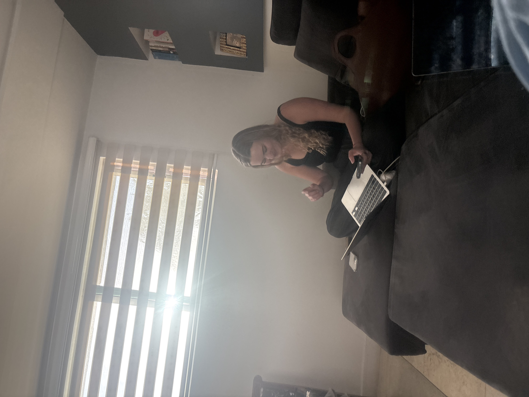 A woman with blonde hair is sitting on a dark gray sofa while working on a silver laptop in a room in San Bernardino Tlaxcalancingo, Mexico. She is wearing a black tank top and black pants. A window with white blinds is to her left. A dark gray wall shelf with books is above her. A dark gray television is on a stand to the right.