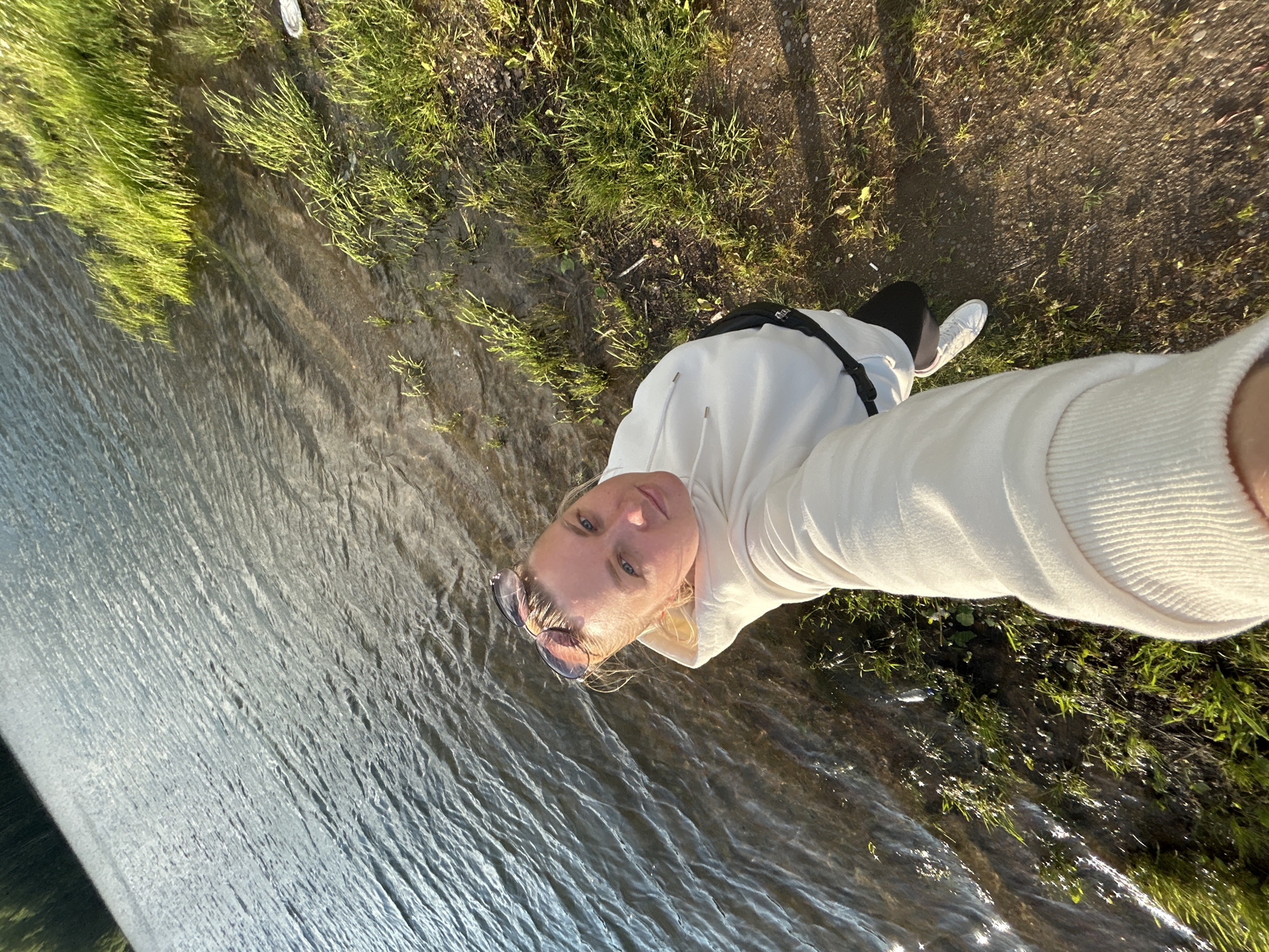 The media file is a selfie taken outdoors during daylight. The image shows a person from the chest up, wearing a white hoodie and black leggings with white sneakers. The person has blonde hair and is wearing sunglasses on their head. They are standing next to a body of water, possibly a river or lake, with green grass and soil visible on the bank. The water appears to be calm with ripples. Sunlight is casting shadows on the ground, suggesting it is either morning or late afternoon. The setting appears to be natural and serene, possibly a park or a natural landscape. There is no visible text in the image.