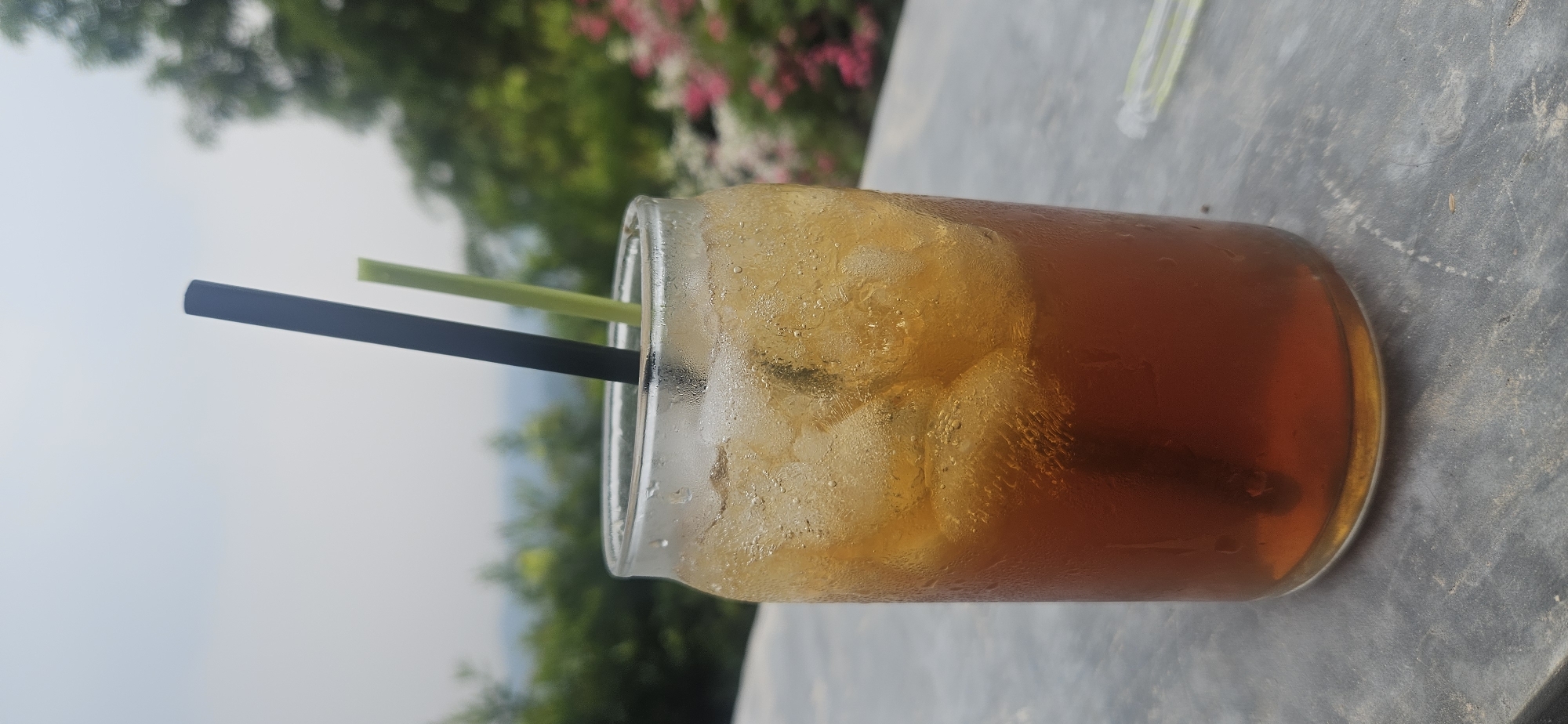 A close-up shot captures a clear glass filled with iced tea, two straws protruding from the drink. The glass is on a textured, gray surface, likely a table. In the background, lush green trees and pink flowers suggest an outdoor setting, possibly a cafe or restaurant in Cijeruk, Indonesia, given the mention of the city. The overall atmosphere is serene, with a focus on the refreshing beverage against a natural backdrop.