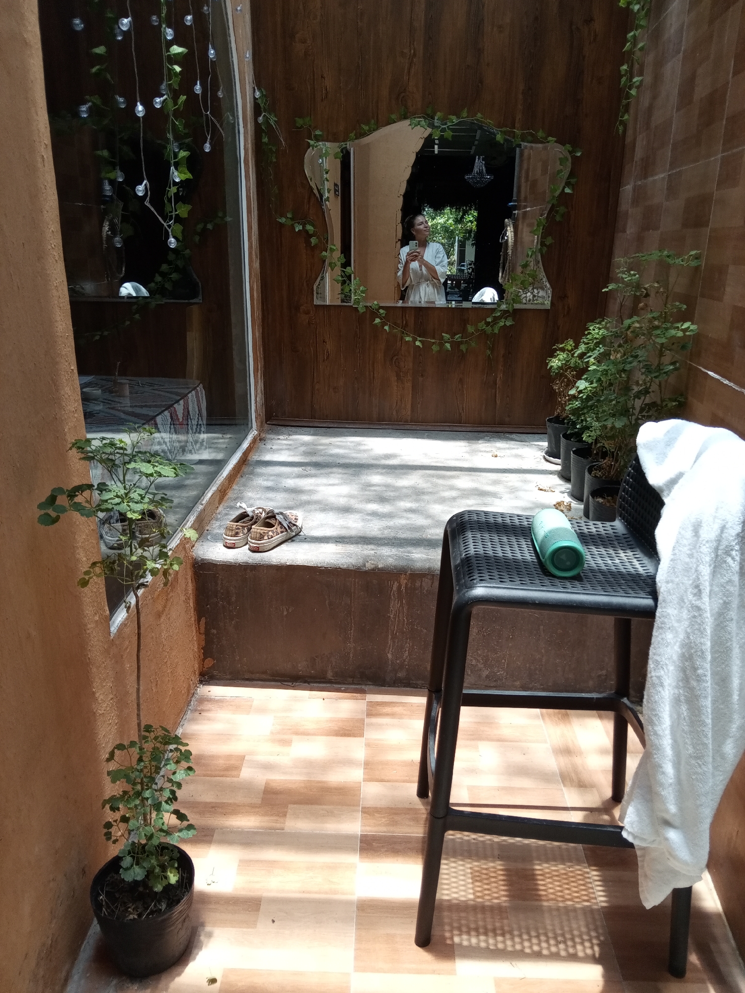 A small, tiled outdoor patio in the Bac Natural Heritage Area Trang, Vietnam, is the scene. The patio features potted plants, decorative lights, and a mirror. A black stool sits in the foreground, upon which rests a green cylindrical object, and draped with a white towel. A pair of sandals rests on the edge of the patio. The mirror reflects a person in a white robe, taking a picture of themselves.