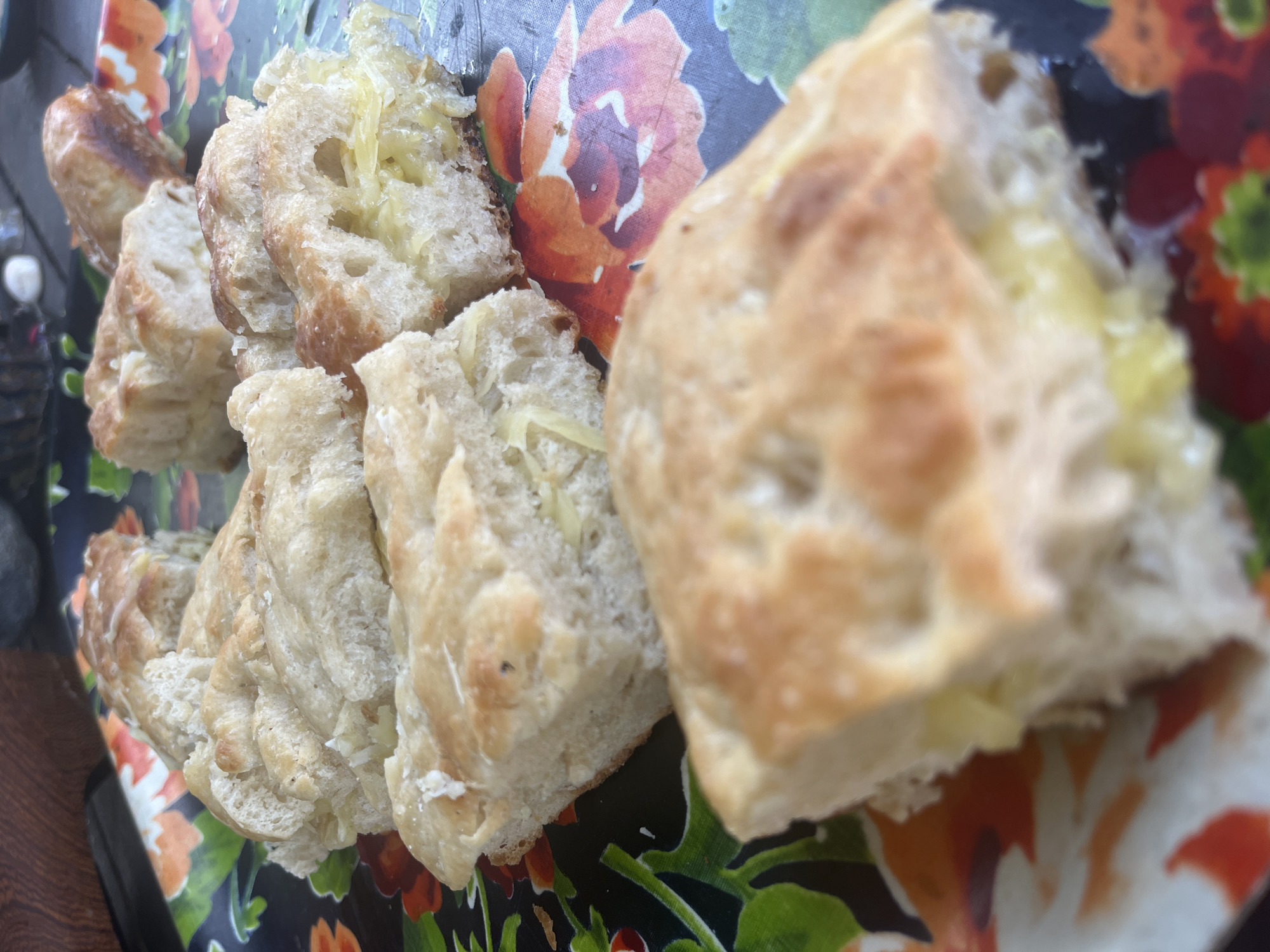 A close-up, overhead shot shows several pieces of sliced focaccia bread arranged on a dark, floral-patterned tray. The bread is golden brown on top with visible dimples and has a light-colored interior, with some pieces revealing a layer of cheese or other filling. The tray has a pattern of large, stylized flowers in shades of orange, red, green, and white. The lighting is natural and appears to be outdoors, casting subtle shadows. The scene is static, with no people or discernible action. The background is blurred, but suggests an outdoor setting with wooden elements and possibly a table.