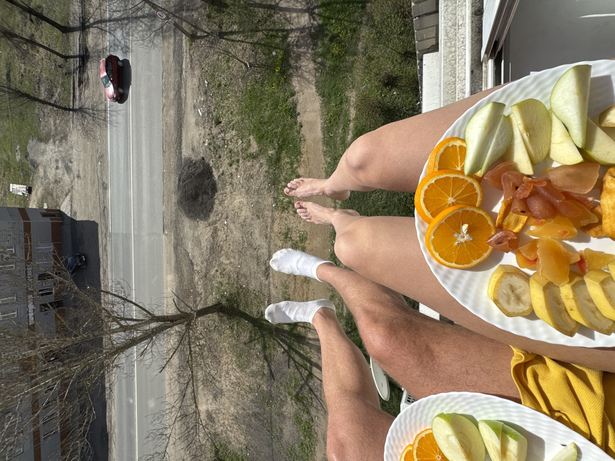 The image captures a high-angle view from a balcony, showcasing the legs of two individuals enjoying a fruit meal, set against an outdoor scene in what appears to be Ново-Никольское, Russia. One person's bare feet are visible, while the other wears white socks. Both are holding or have resting on their laps white plates filled with sliced fresh fruit, including oranges, green apples, and bananas, along with various types of dried fruit. A yellow fabric item, possibly a towel or napkin, is also visible.
Below the balcony, a paved road with clear white lane markings stretches out. A red car is visible on the road, with another dark car partially seen near a grey multi-story building. This building has visible windows and text partially reading "PROPERTIES" on its facade. Bare trees, casting long shadows across the ground, line the roadside, suggesting strong sunlight, indicative of either morning or late afternoon. The ground is a mix of dry grass and dirt. A traffic sign with a red circle and the number "30" is visible near the red car. The weather is sunny and clear.