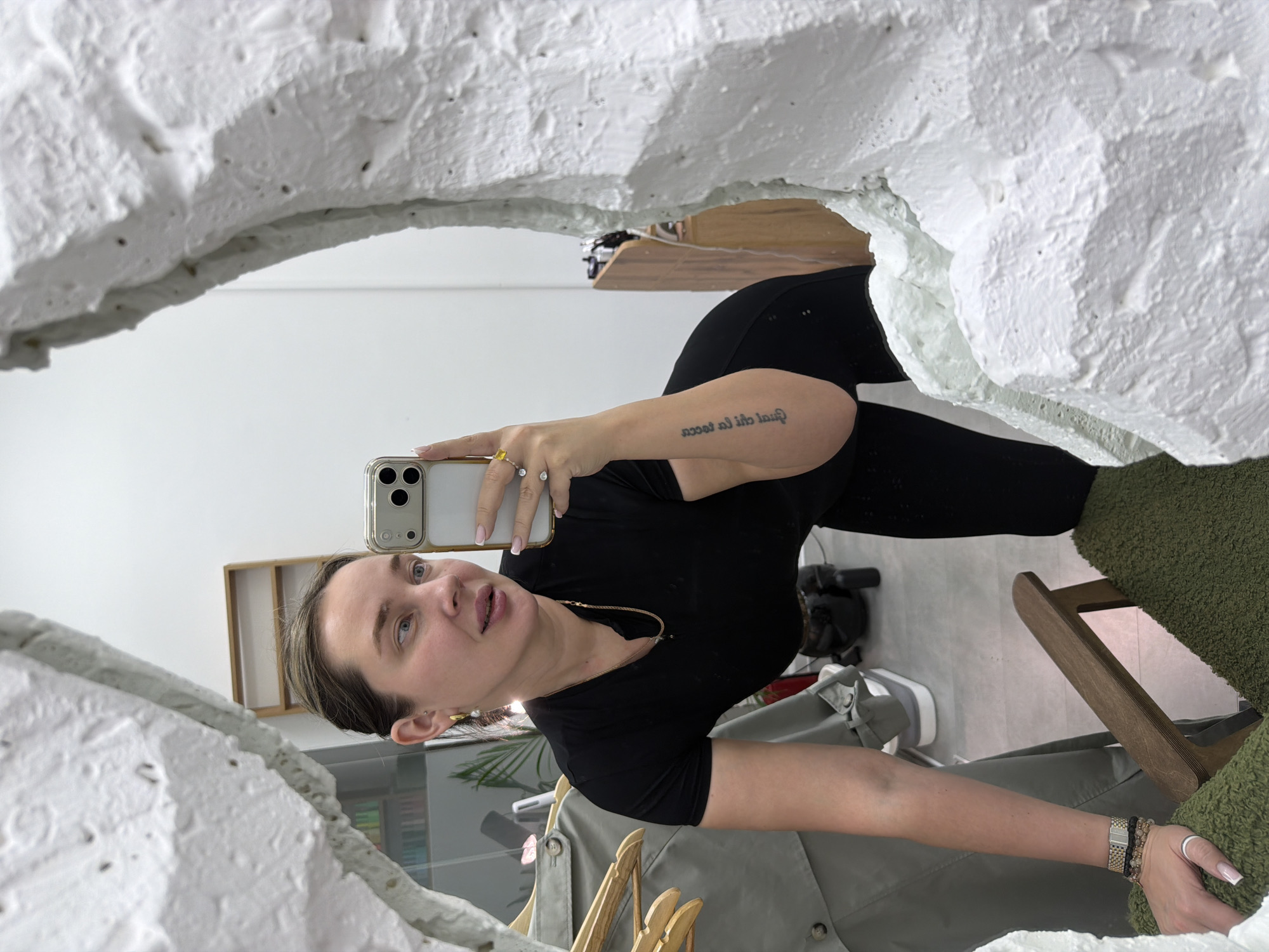 A woman is taking a selfie in a mirror with a textured white frame. She is wearing a black top and black leggings. She is holding a smartphone in her right hand. Her arm is visible, and there is a tattoo on her inner forearm. She is wearing rings on her fingers and a bracelet on her left wrist. A wooden chair with a green cushion is visible behind her. In the background, there are clothes hanging, a gray jacket, shelves, a cabinet, a box, a vacuum cleaner, and a pair of white shoes. The setting appears to be inside a room with white walls. There is no explicit information to indicate the location of this image in Moscow, Russia.
