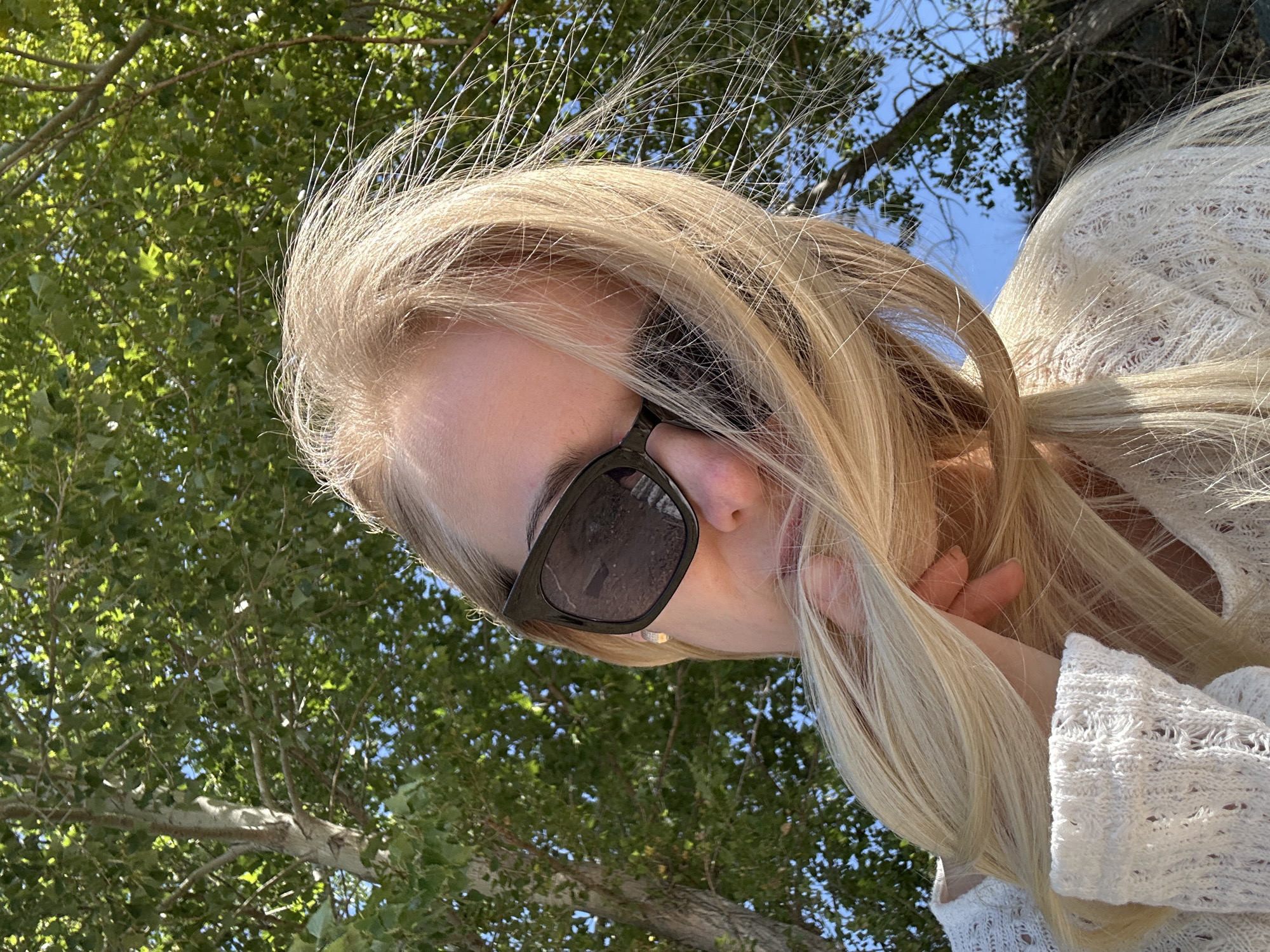 The image shows a woman with long blonde hair and sunglasses, wearing a white knitted sweater, outdoors. Her hair is blowing in the wind, partially obscuring her face. She is holding her hair with her left hand. The background consists of trees and a blue sky. The location is specified as Gornyi Erik, Russia.