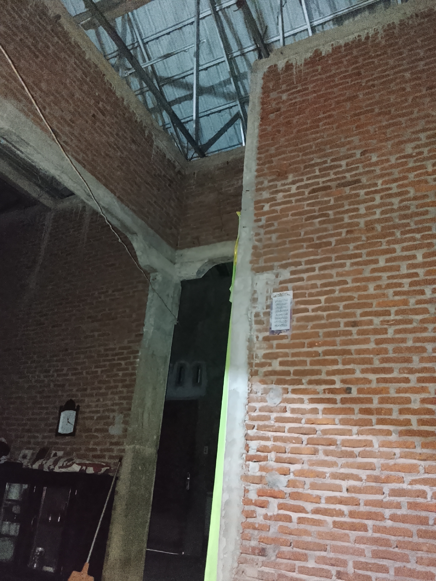 The image depicts an interior scene of a building under construction or renovation in Tanjongbabahkrueng, Indonesia. The walls are made of exposed brick, with some sections plastered with concrete. A metal roof structure is visible overhead. In the foreground, a large concrete pillar divides the space. To the left, a clock is mounted on the brick wall above a dark wooden cabinet. A broom leans against the wall. The background shows an open doorway leading to a darker area, with glimpses of what appear to be interior doors or walls. A light green tarp or material is present along the edge of the pillar on the right. A small piece of paper, possibly a notice or calendar, is affixed to the brick wall on the right side of the image. The scene appears to be in a state of transition, with unfinished elements and a sense of domestic utility.