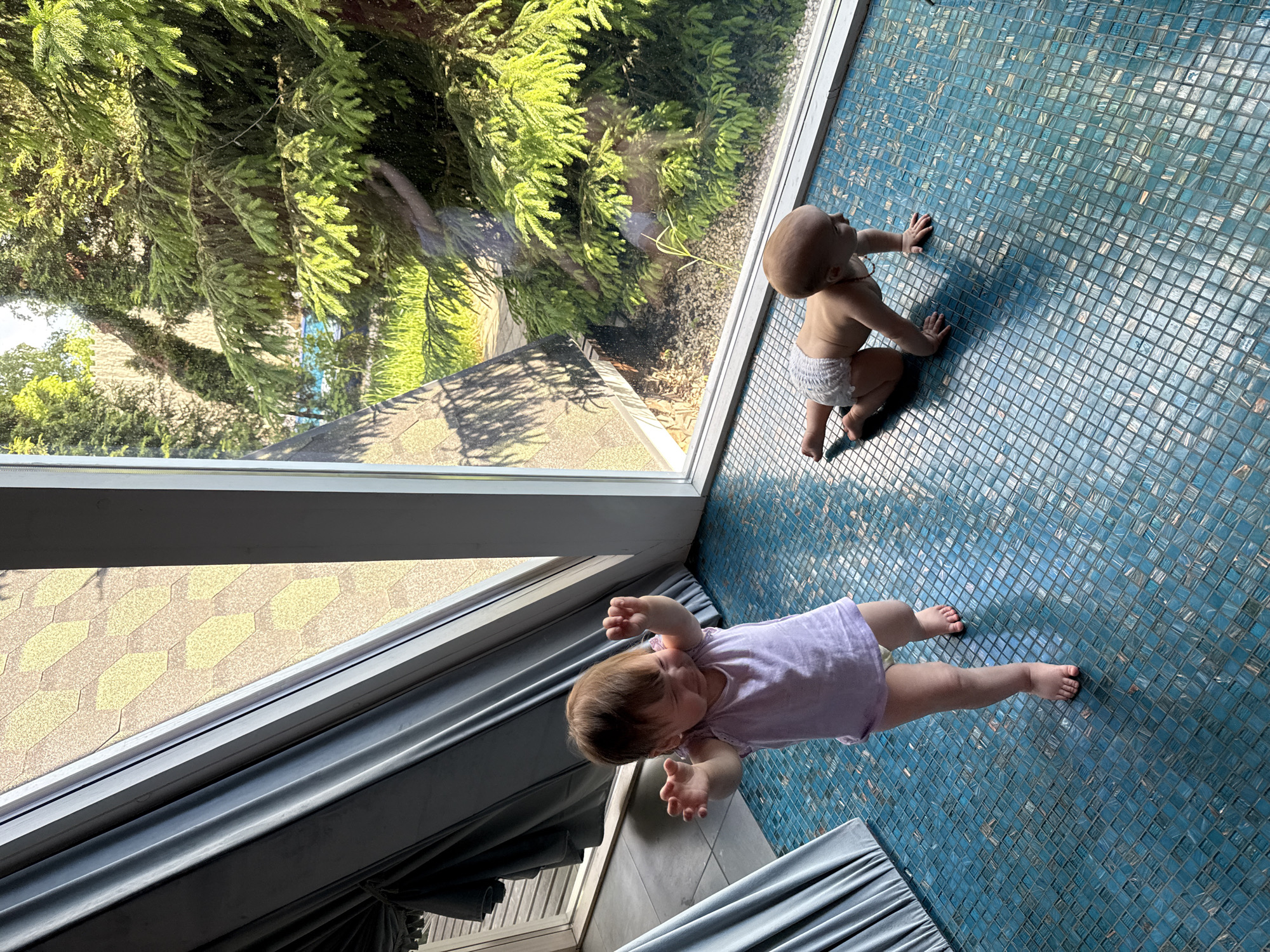 Two infants are on a tiled floor indoors near a window. One infant wears a light purple shirt and is lying on their stomach. The other infant wears a diaper and is on their hands and knees. Outside the window, there are trees and a building with a tan roof. The specific location in the city of Чёрное, Russia is impossible to ascertain from the visual information.