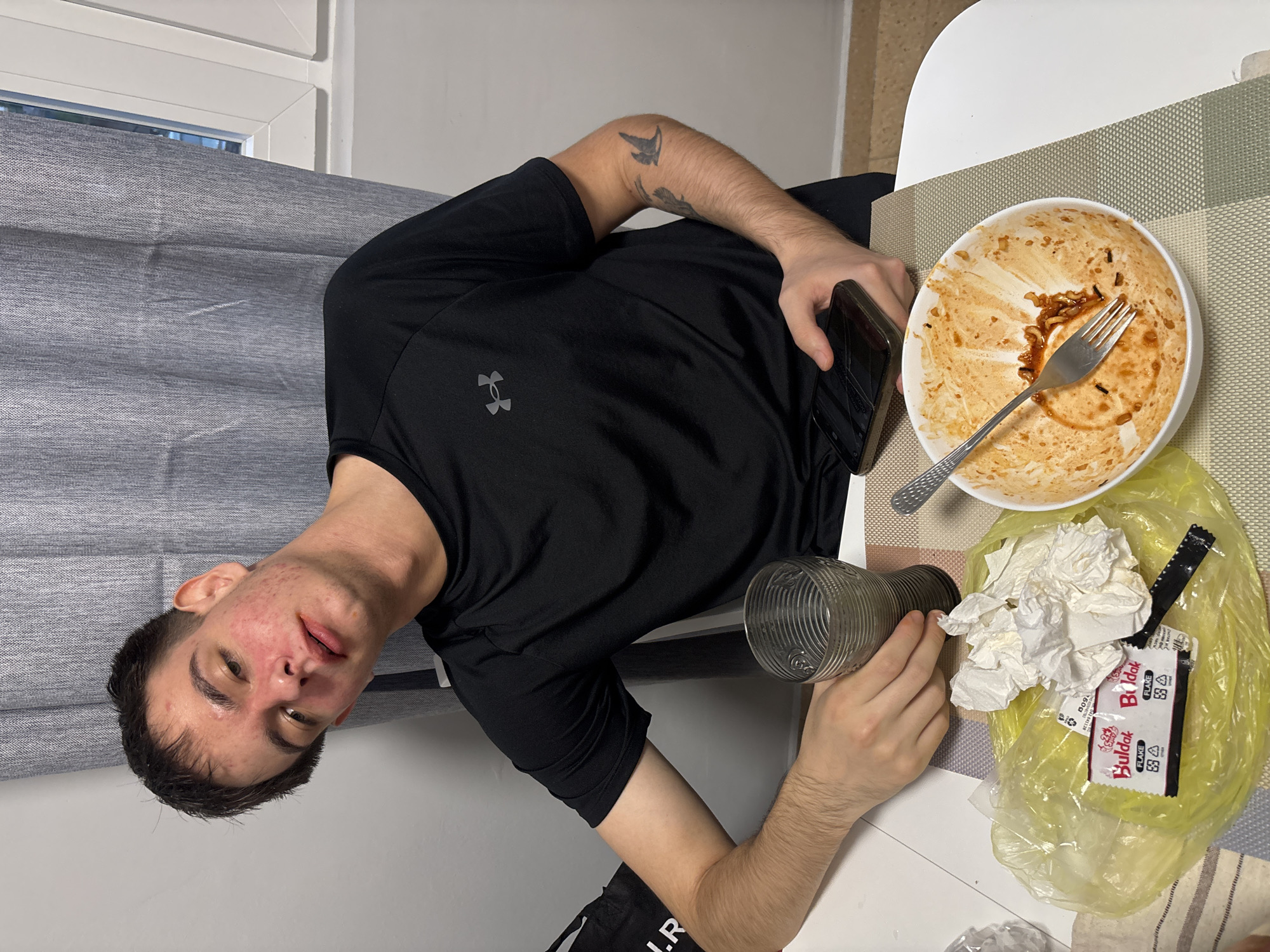 A young man is seated at a table indoors, possibly in Plovdiv, Bulgaria, appearing to have recently finished a meal. He is oriented in a way that suggests the photo was taken from above and to his left, with his head turned towards the camera. He wears a black short-sleeved t-shirt with a grey Under Armour logo on the chest. His face shows some visible acne, and his dark hair is short. On his right upper arm, a black tattoo with abstract shapes or perhaps birds is visible.
He holds a smartphone with a cracked screen in his left hand, and a clear, ribbed glass in his right hand. The table is white, and a light-colored woven placemat is underneath a dirty white bowl with a metal fork resting in it, suggesting remnants of noodles or ramen. Next to the bowl is a crumpled pile of white napkins and a yellow plastic bag. Inside the bag, a "Buldak FLAKE" packet is visible, along with a recycling symbol and the word "RECYCLABLE." The packet also shows "ДОЗА" (Doza) and possibly "БЪЛДЪК" (Buldak in Bulgarian).
In the background, a grey curtain or blind covers part of a window, through which some natural light can be seen. The overall setting is simple and domestic. The man's expression is neutral.