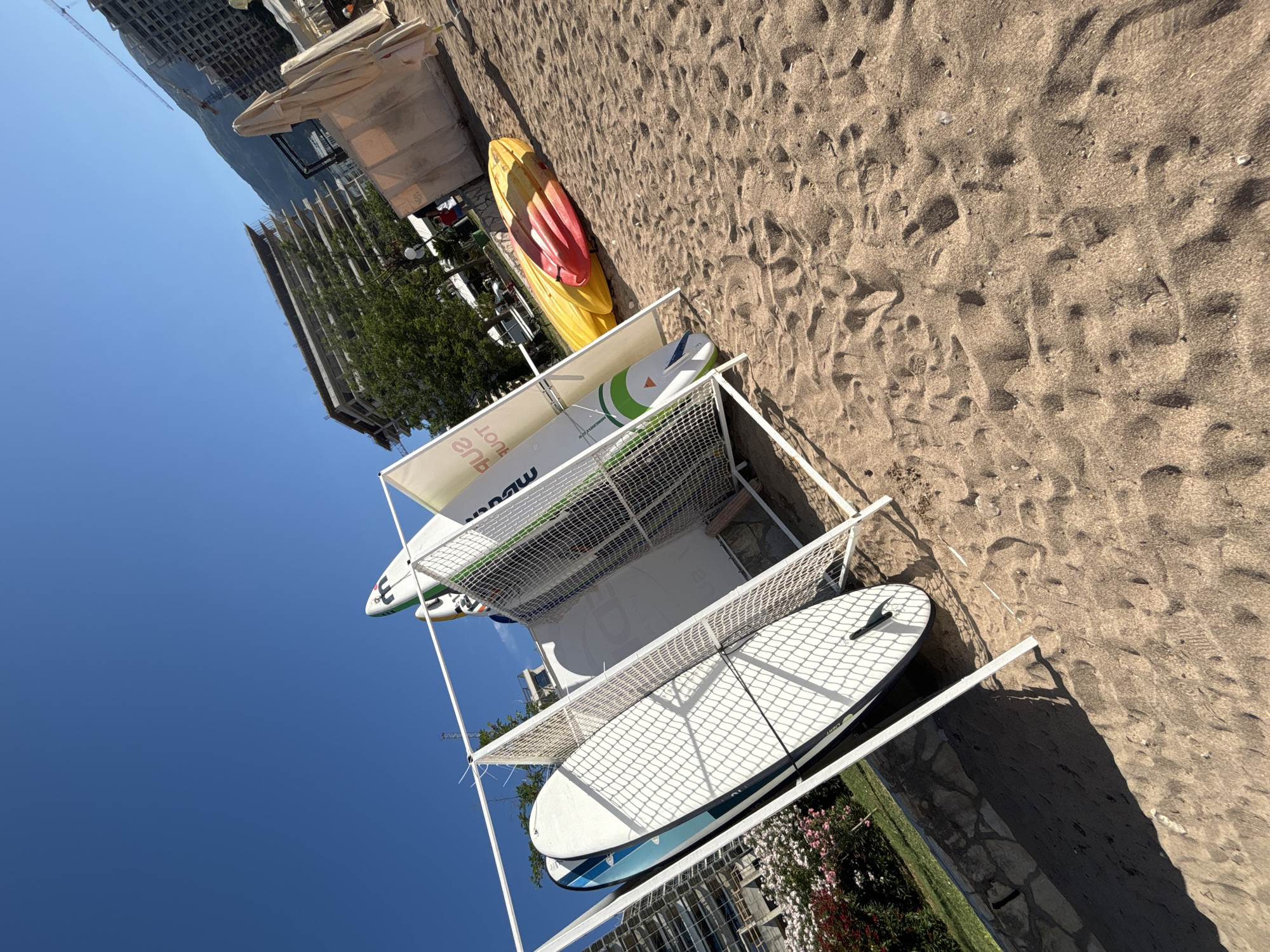 On a sunny day in Budva, Montenegro, a beachside scene is captured. The foreground is dominated by sand, with subtle textures and shadows suggesting the uneven surface. In the midground, a white metal rack holds several paddleboards upright. One paddleboard, on the left, is white with blue accents and a prominent fin. Behind it, another paddleboard is partially visible, with a green and white design. To the right of the rack, two kayaks, one red and one yellow, are resting on the sand. Beyond the kayaks, large, folded beige beach umbrellas stand, and in the far background, unfinished high-rise buildings under construction are visible against a clear blue sky, with a mountainside visible to the right. A small patch of green foliage with pink flowers is seen to the left of the paddleboard rack.