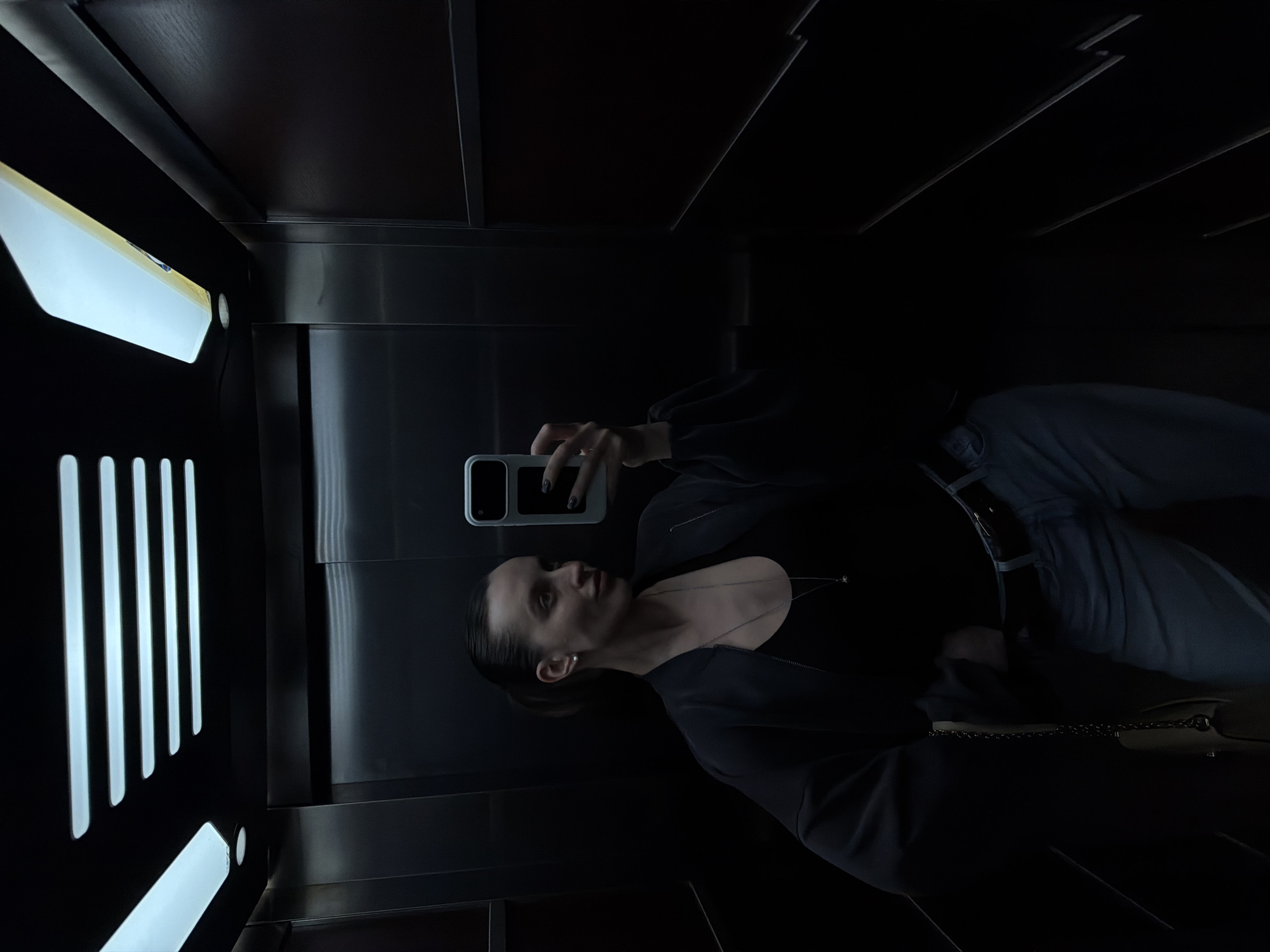 An individual, likely female, takes a selfie in what appears to be an elevator or a confined, reflective space. The person is oriented sideways in the image, suggesting a mirror reflection or a camera held to capture the reflection. She has dark hair pulled back, wears a dark long-sleeved top or jacket, light-colored trousers, a delicate necklace with a pendant, and small pearl earrings.
She holds a light-colored smartphone with a dual-camera setup in her right hand, positioned to capture the image. A light-colored handbag with a chain strap is visible on her left side. The setting features dark, possibly metallic or wood-paneled walls that reflect the bright, elongated rectangular light fixtures positioned on the left side of the frame. These lights create strong illumination on one side and deep shadows across much of the environment. The overall lighting is dim, emphasizing the contrast between the illuminated areas and the dark surfaces. The person's expression is neutral as she looks towards the phone. No text is visible. The scene is located in Budva, Montenegro.