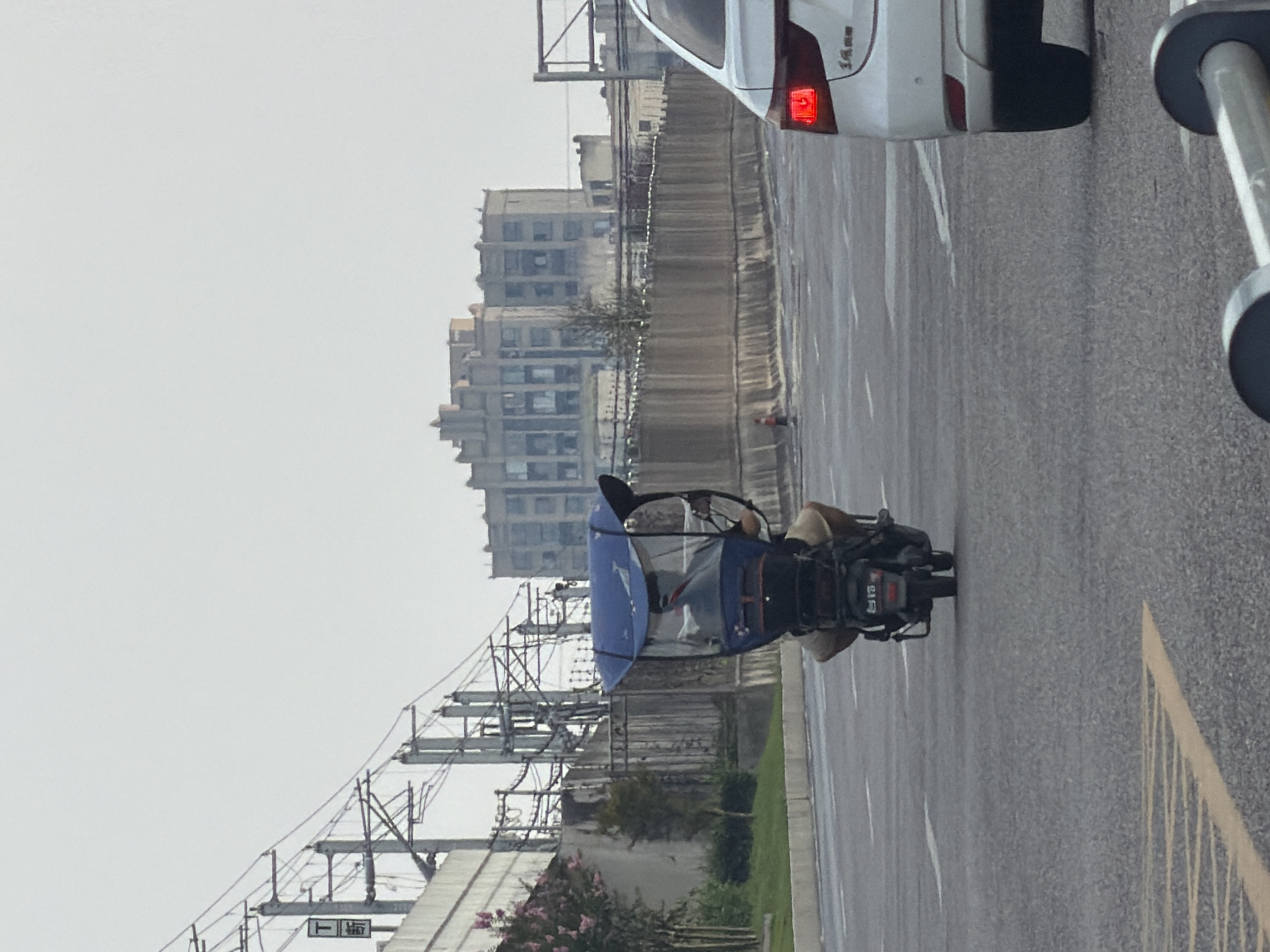 A rear view shows a person riding a three-wheeled electric scooter on a paved road in Jiangjin, China. The scooter is equipped with a blue canopy that covers the rider and a portion of the rear. The rider is wearing light-colored pants and a tan shirt. The scooter has a license plate with Chinese characters and numbers. In the foreground, a white car is visible on the right, with its red taillight illuminated. In the background, a long, textured wall runs parallel to the road. Beyond the wall, a multi-story building with a unique, stepped architectural style is visible against an overcast sky. Overhead, power lines and poles are present, indicating urban infrastructure. The scene appears to be captured during daylight hours.