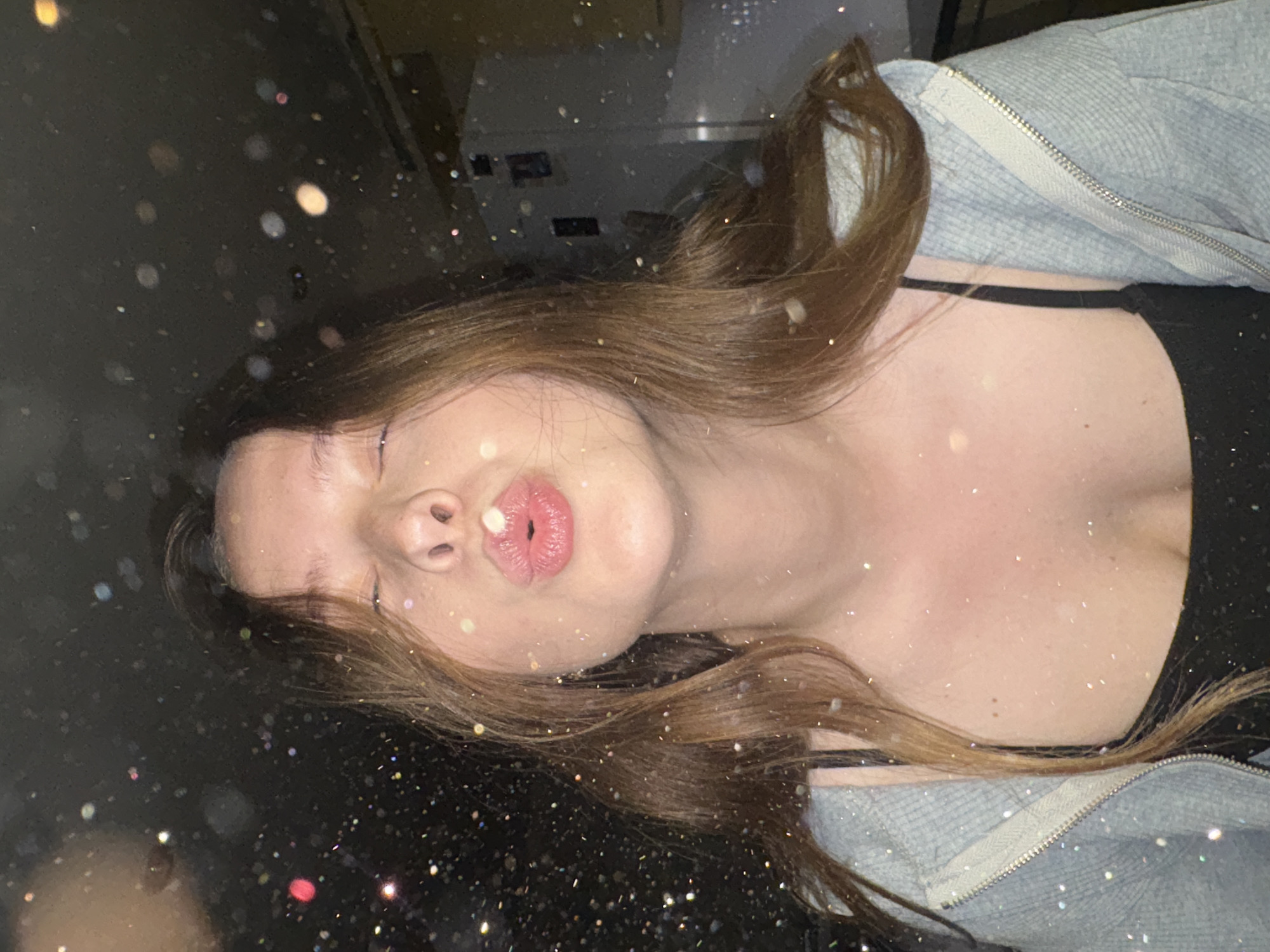 The image shows a young woman with long brown hair, her eyes closed, and her lips pursed in a kissing gesture. She is wearing a black tank top and a light blue zippered hoodie. The background is dark and appears to be indoors, possibly a kitchen, with a white refrigerator visible on the right. There are numerous out-of-focus lights or glitter particles scattered across the image, creating a bokeh effect. The location is identified as Chaikovsky, Russia.