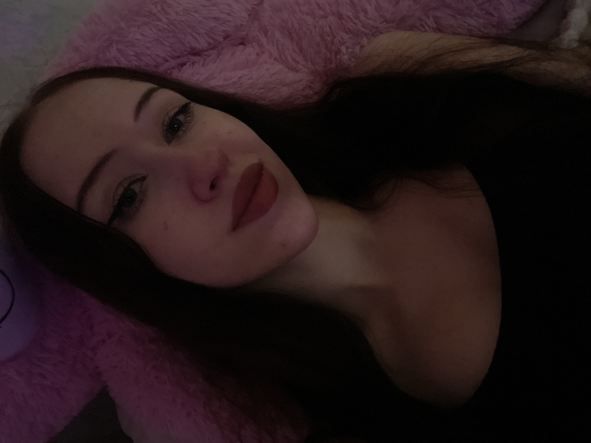 A young woman with long dark hair and dark-colored clothing is resting on a pink, fluffy surface, possibly a large stuffed animal or blanket. She is looking towards the camera, with her makeup including eyeliner and lipstick. The lighting is dim, with a pinkish hue casting over the scene. No specific location in Votkinsk, Russia, can be determined from the image.