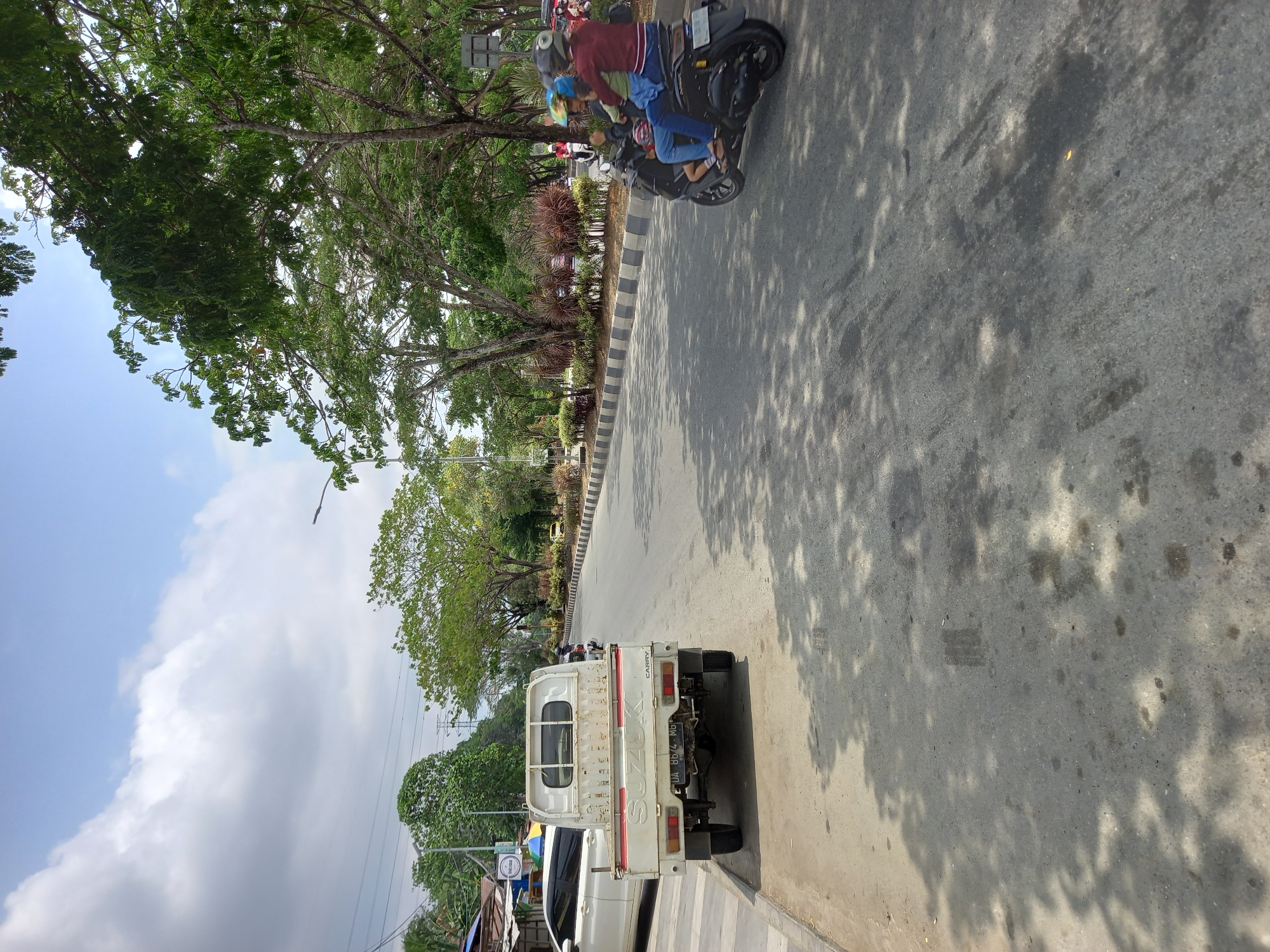 A sunny day with patchy clouds is depicted on a road in Samarinda, Indonesia. A white Suzuki Carry pickup truck is parked on the left side of the road, adjacent to a sidewalk and some buildings. A white car is parked in front of the truck. Further down the road, a scooter carrying three people, two adults and a child, is traveling away from the camera. The road is lined with trees and a median strip with decorative plants. The scene is illuminated by sunlight, casting shadows from the trees onto the road surface.