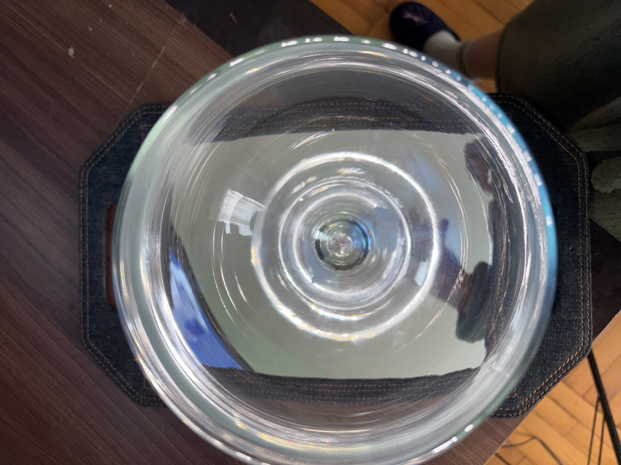 A clear glass bowl sits on a dark denim coaster. The coaster is placed on a wooden surface. A person wearing a slipper is visible near the bowl. The reflection in the bowl shows the environment. The city and country are unknown.